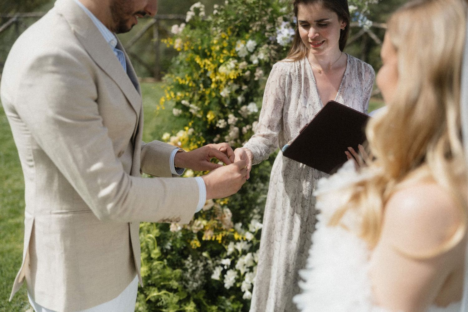 Groom placing a wedding ring on the bride's finger during an outdoor ceremony with a floral arch backdrop.