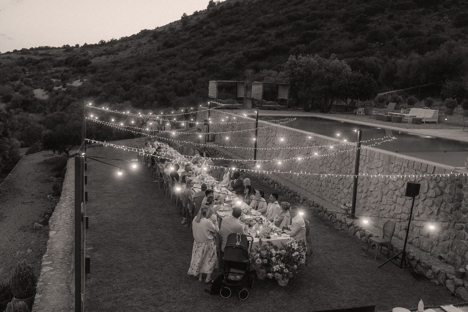 Black and white aerial view of an elegant outdoor dinner party with string lights along a long table at dusk.