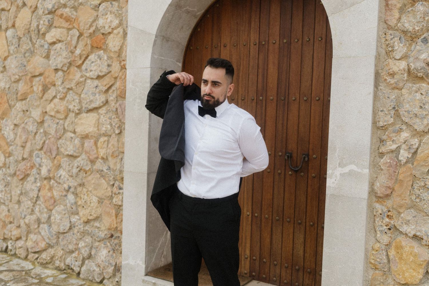 Stylish groom in black tuxedo with bow tie holding jacket over shoulder near stone archway.