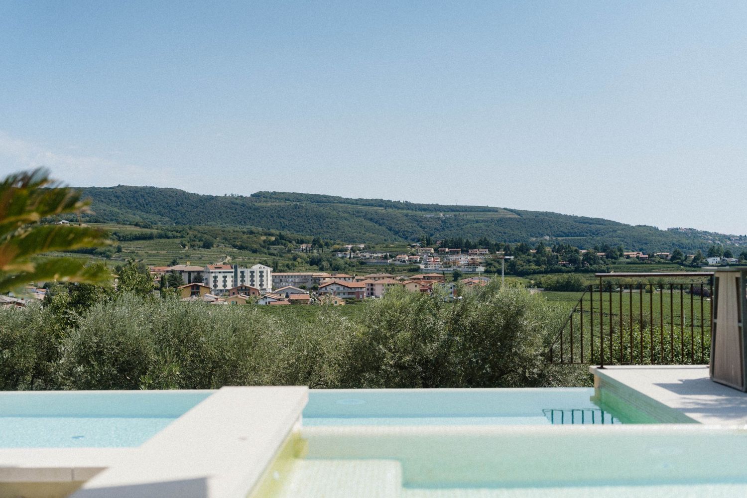 Infinity pool overlooking lush olive trees and a scenic hilltop village under a clear blue sky.
