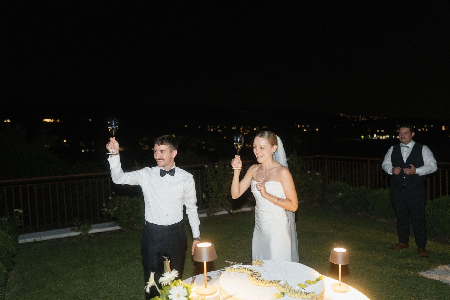Bride and groom raising champagne glasses at night by their wedding cake during an outdoor evening celebration.