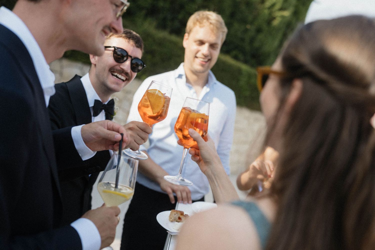 Group of elegantly dressed people toasting with champagne and Aperol Spritz at an outdoor wedding reception.