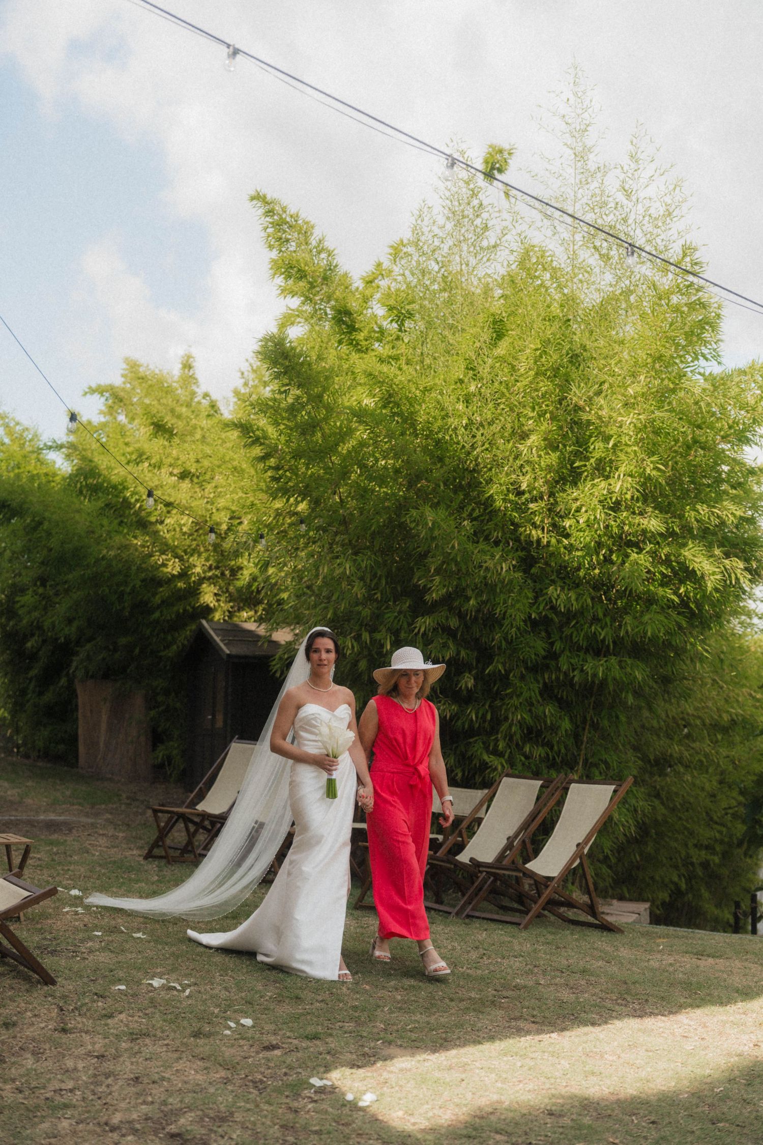 Bride in white gown with veil walks with woman in red dress at outdoor wedding.