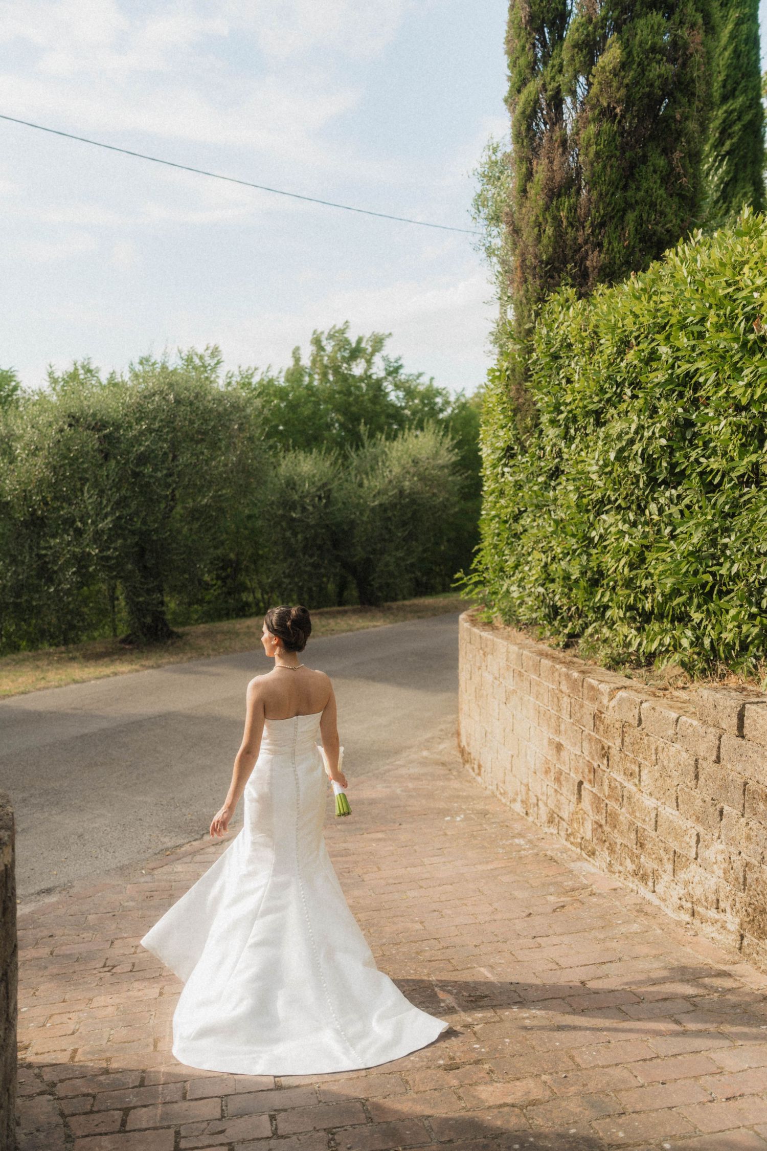 Bride in white strapless gown walking down a rustic stone path lined with lush green hedges and trees.