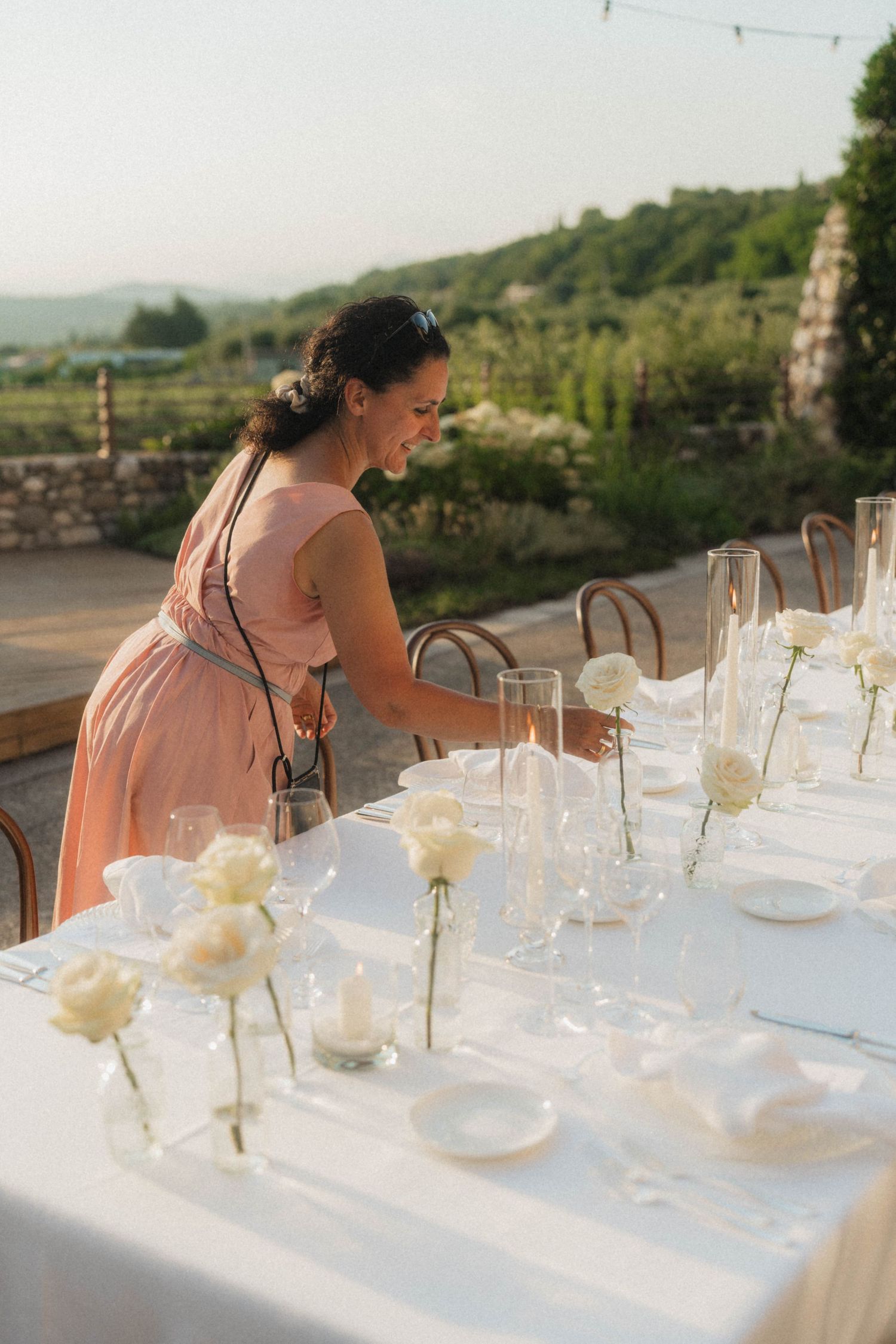 Woman in pink dress arranging white roses and candles on elegant outdoor wedding table.