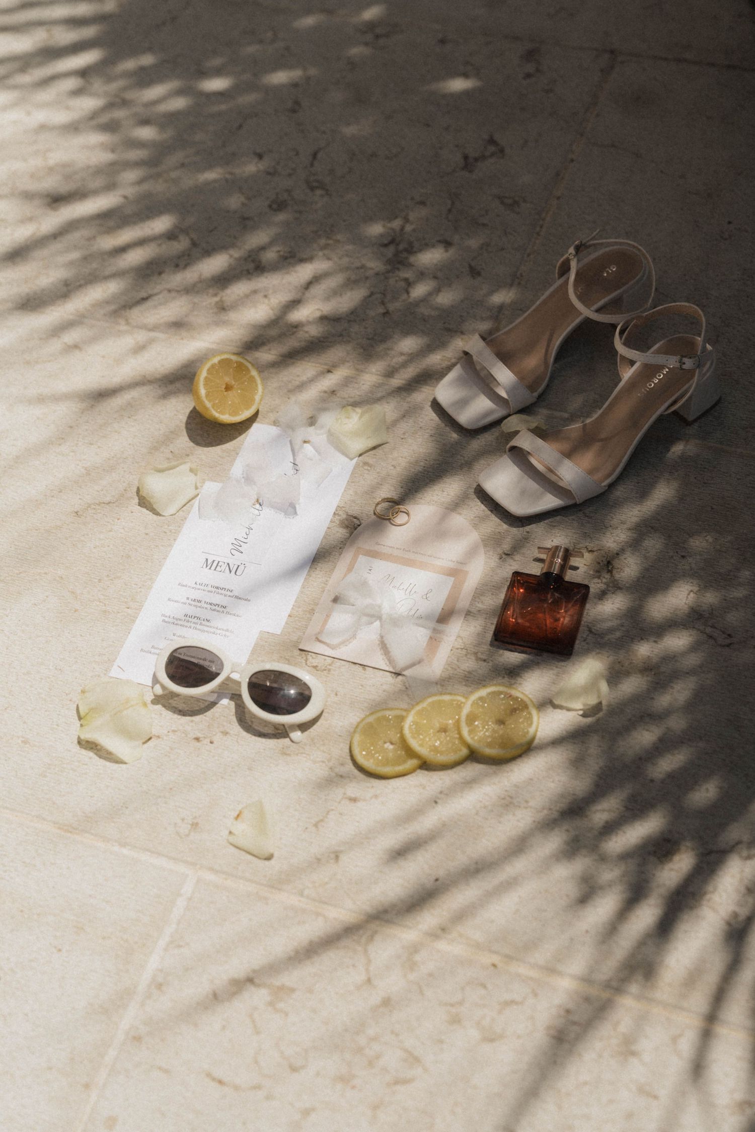 Elegant wedding flat lay with nude heels, perfume, sunglasses, lemon slices, and floral petals on marble floor.