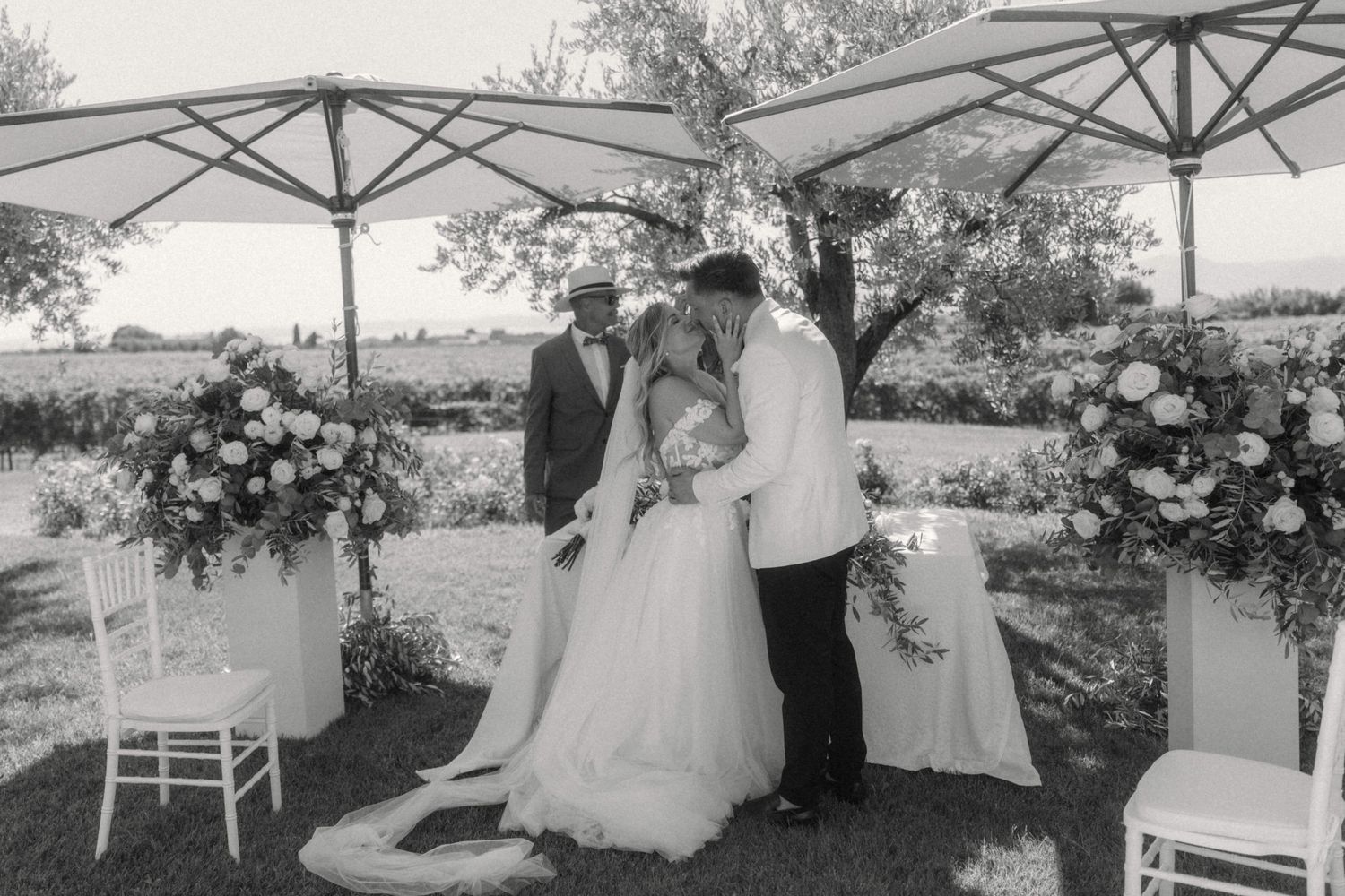 Bride and groom share a kiss at outdoor wedding ceremony with floral arrangements in black and white photo.