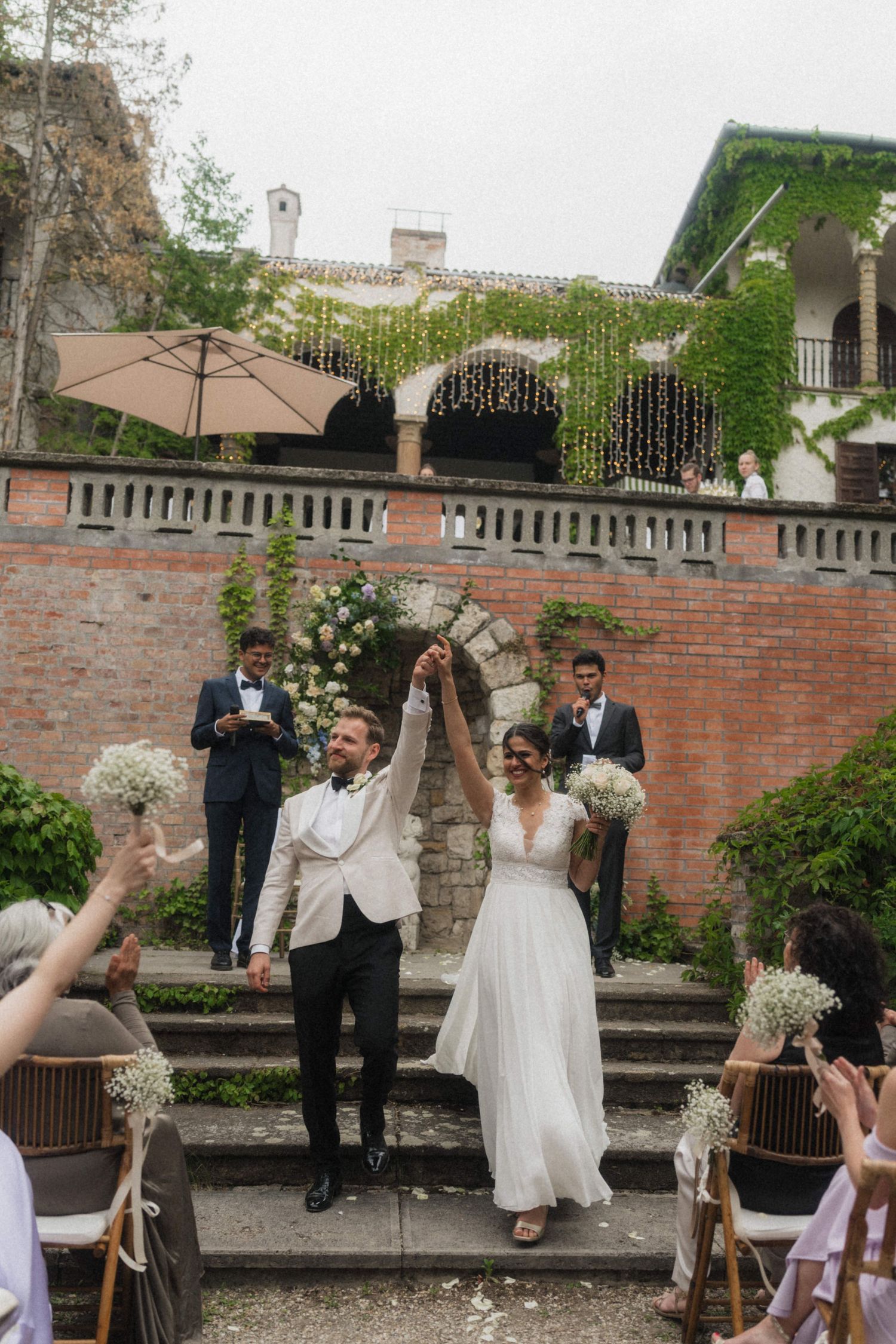 Joyful bride and groom raising hands after outdoor wedding ceremony at ivy-covered venue.