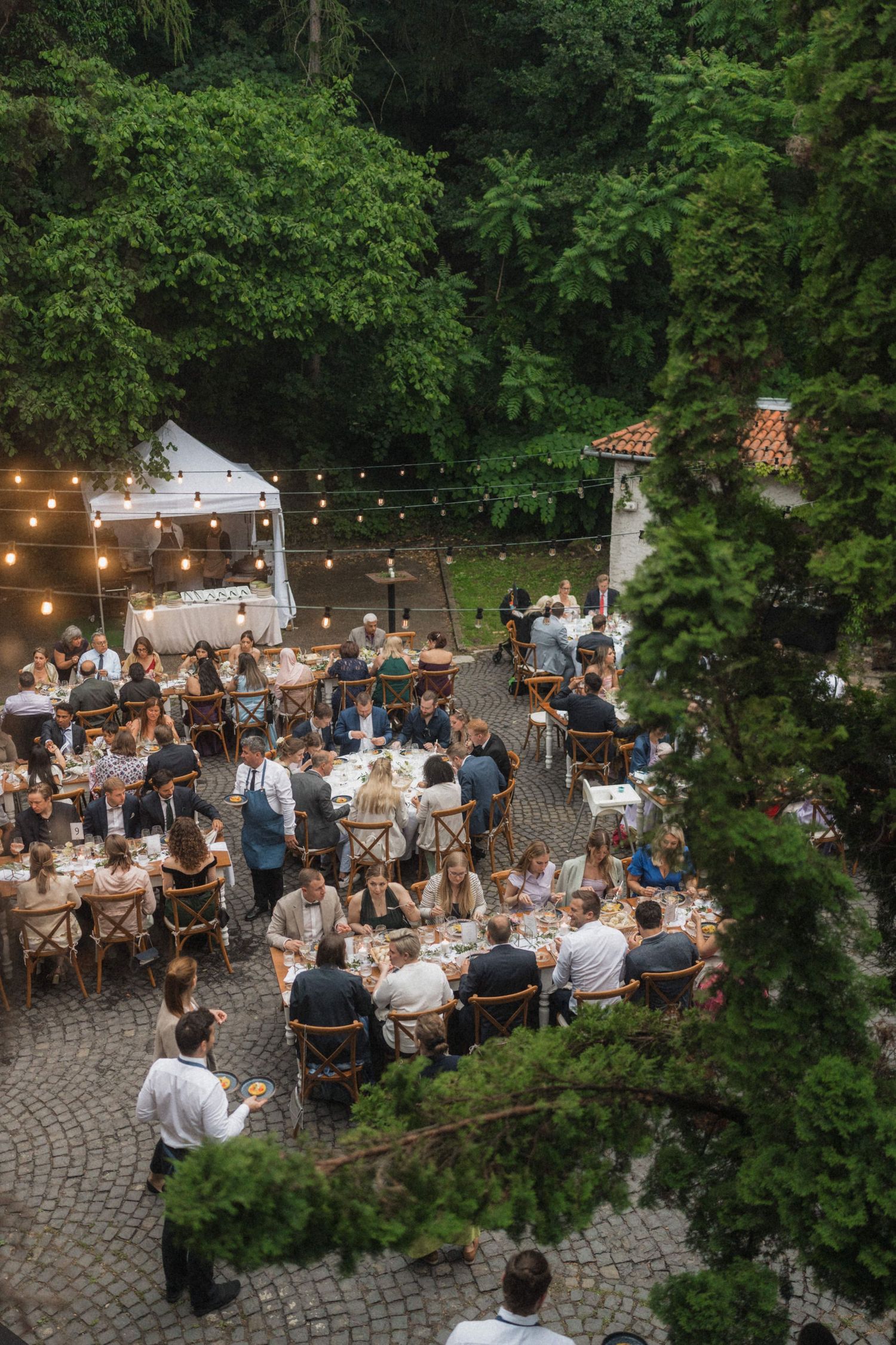 Aerial view of elegant outdoor wedding reception with round tables and string lights in garden.