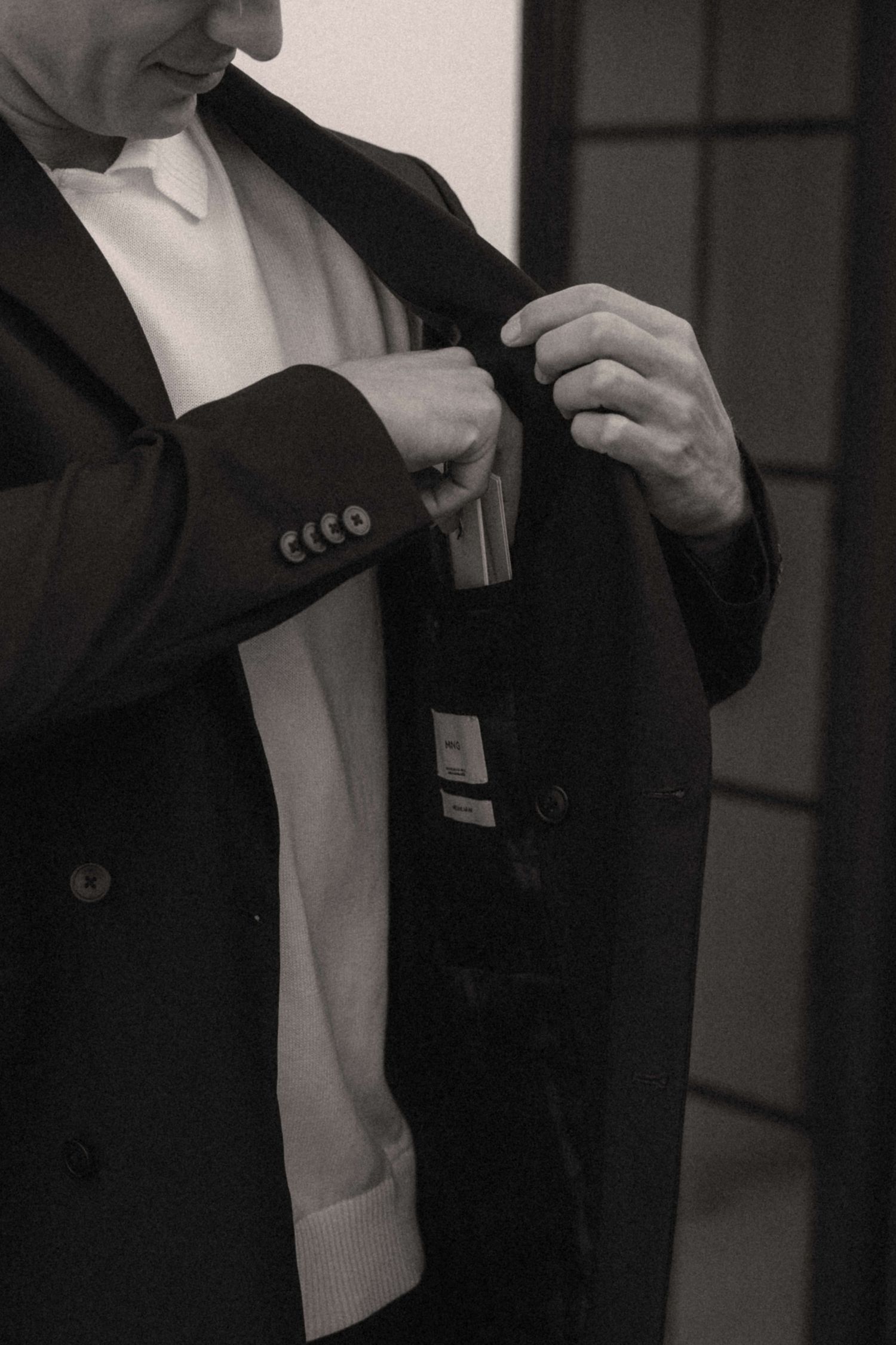 Black and white photo of a groom adjusting his dark double-breasted suit jacket before the wedding ceremony.