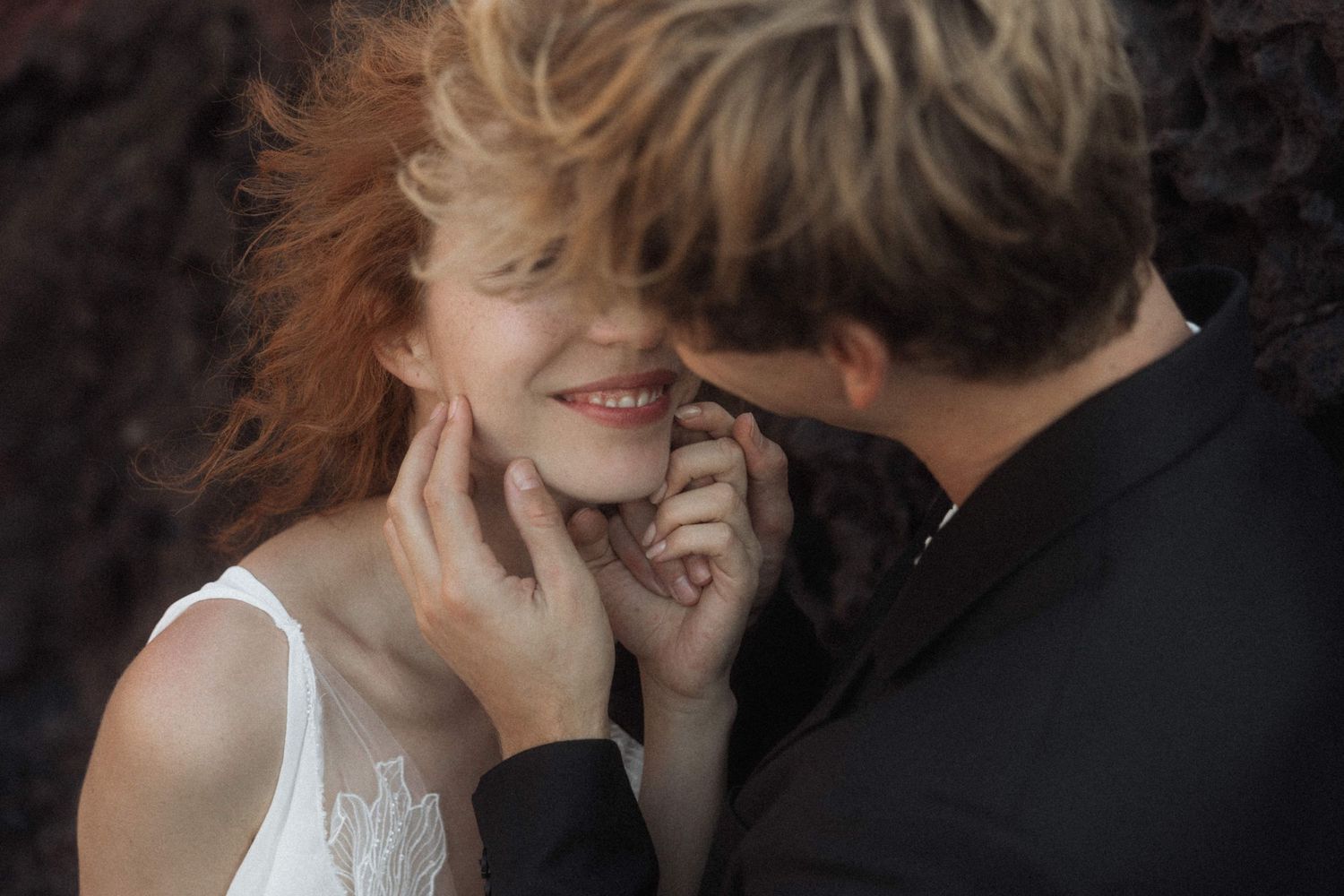 Groom tenderly holds brides face as she smiles joyfully against a dark volcanic rock backdrop.