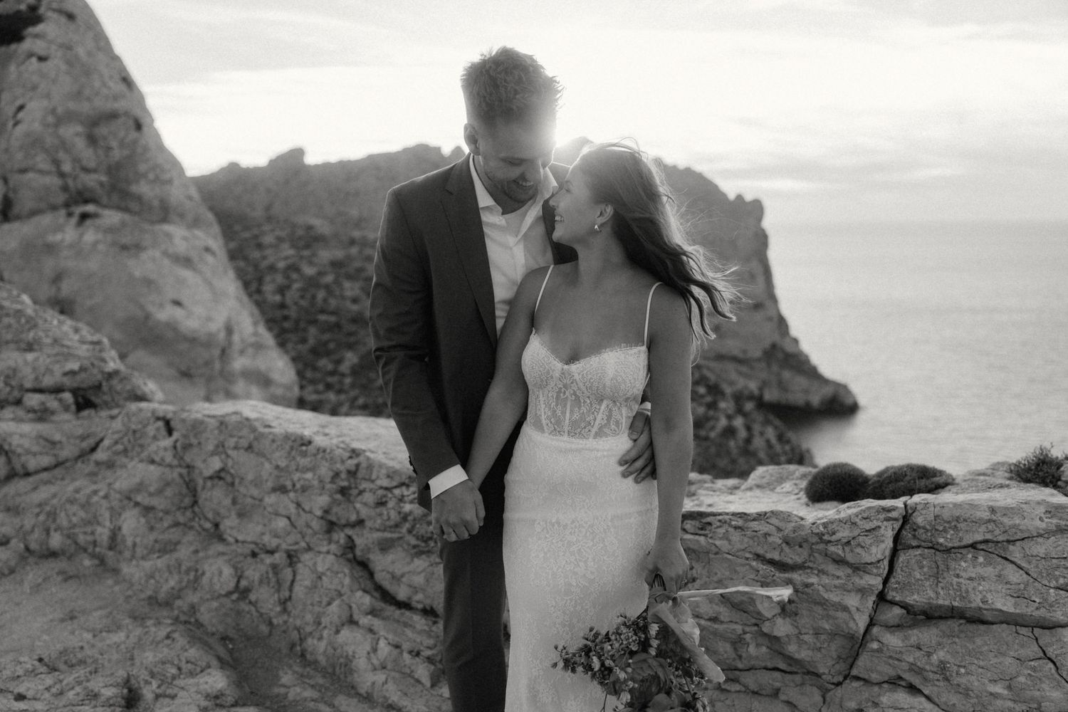 Black and white photo of a bride and groom holding hands on rocky cliffs by the ocean at sunset.