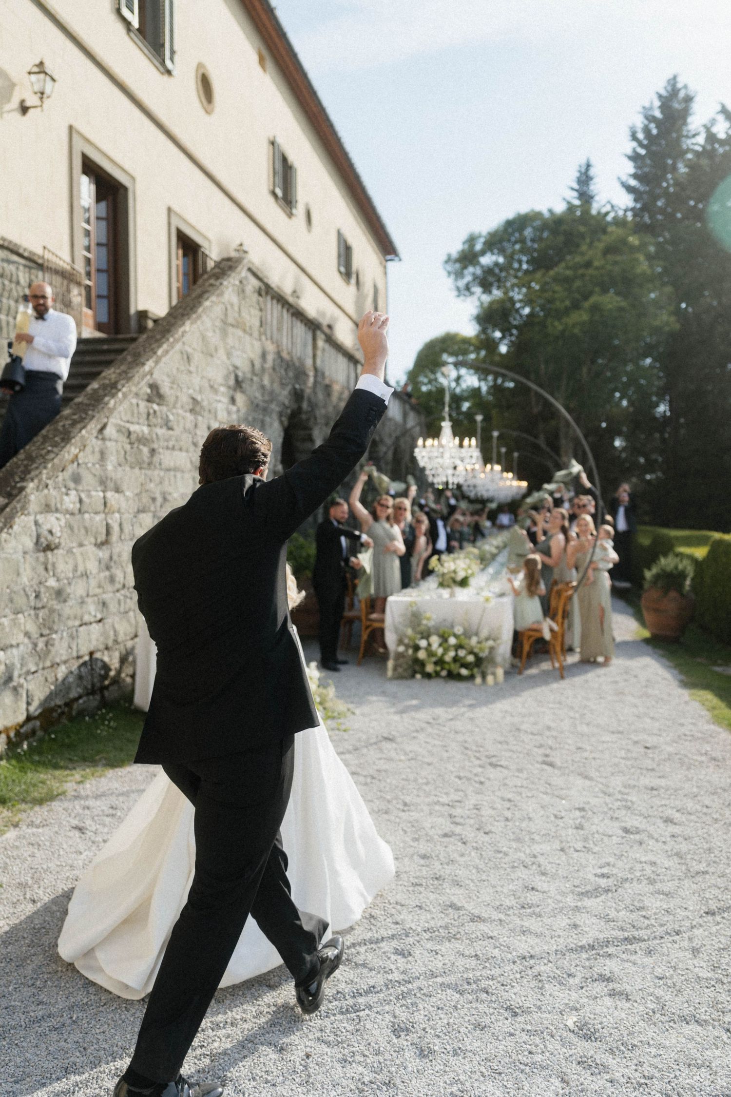 Groom raises hand triumphantly leading bride in white gown to outdoor wedding reception at Italian villa.