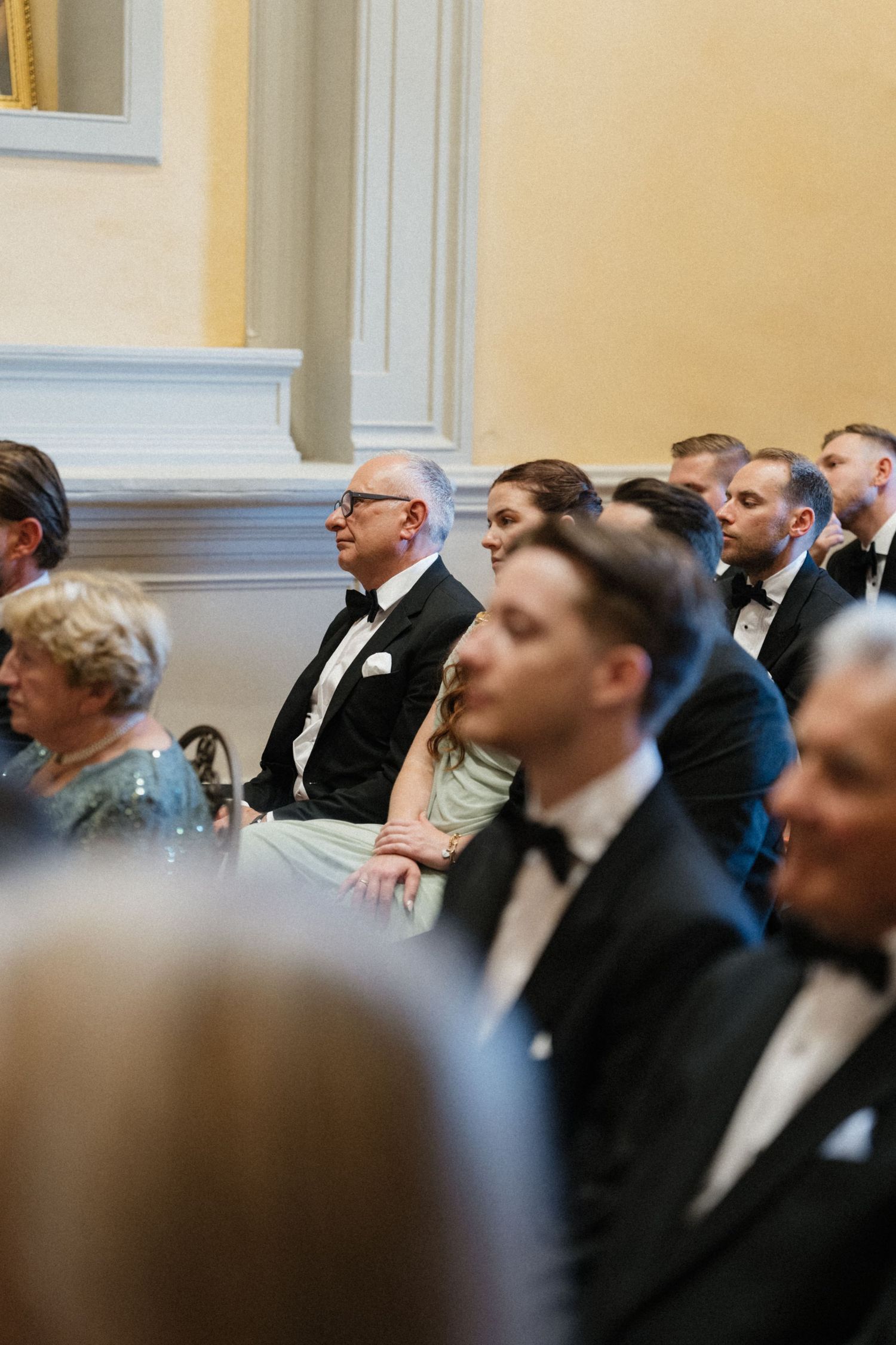 Elegantly dressed wedding guests seated in golden-walled church attentively watching the ceremony.
