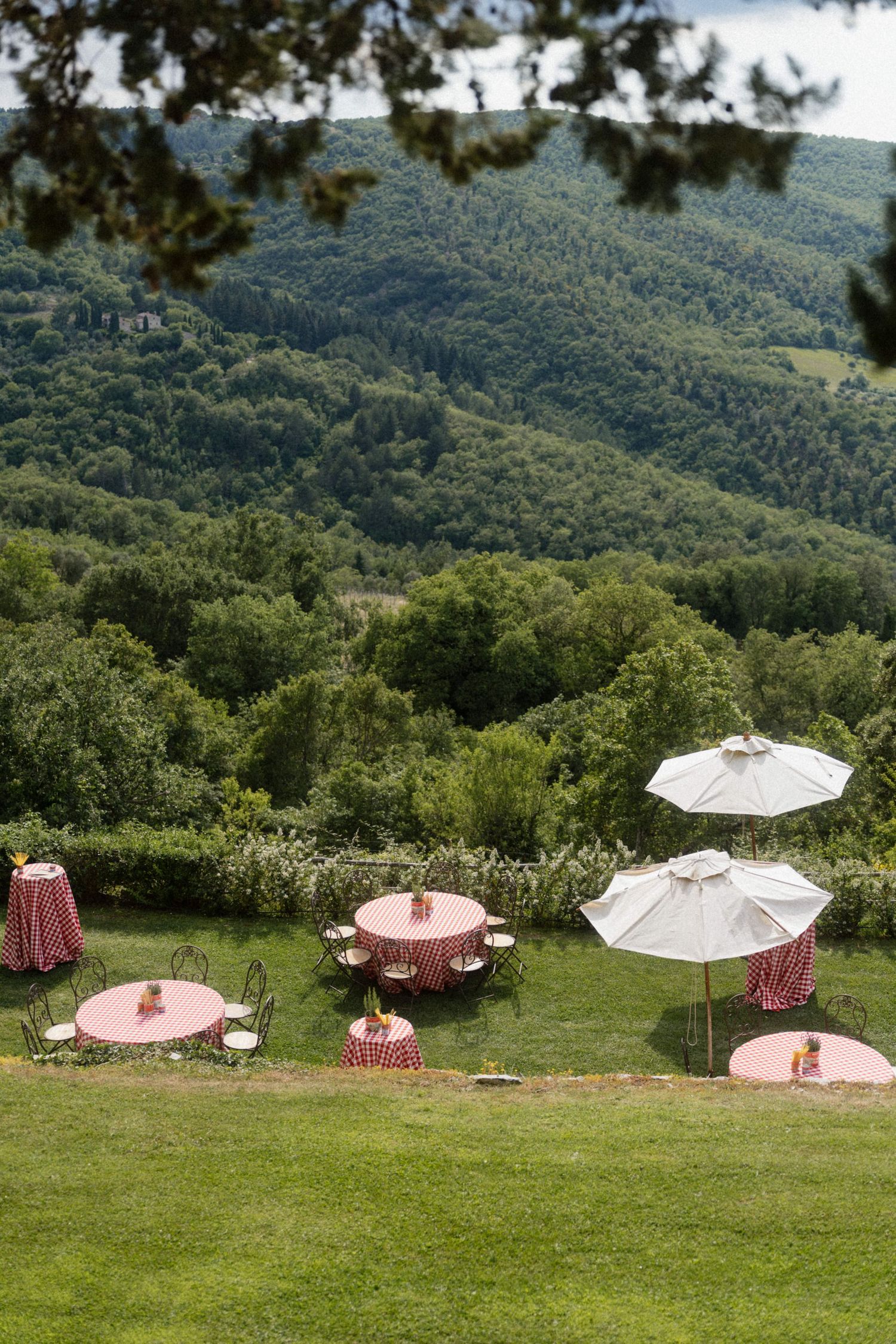 Elegant outdoor wedding reception tables with red checkered cloths and white umbrellas overlooking lush green hills.