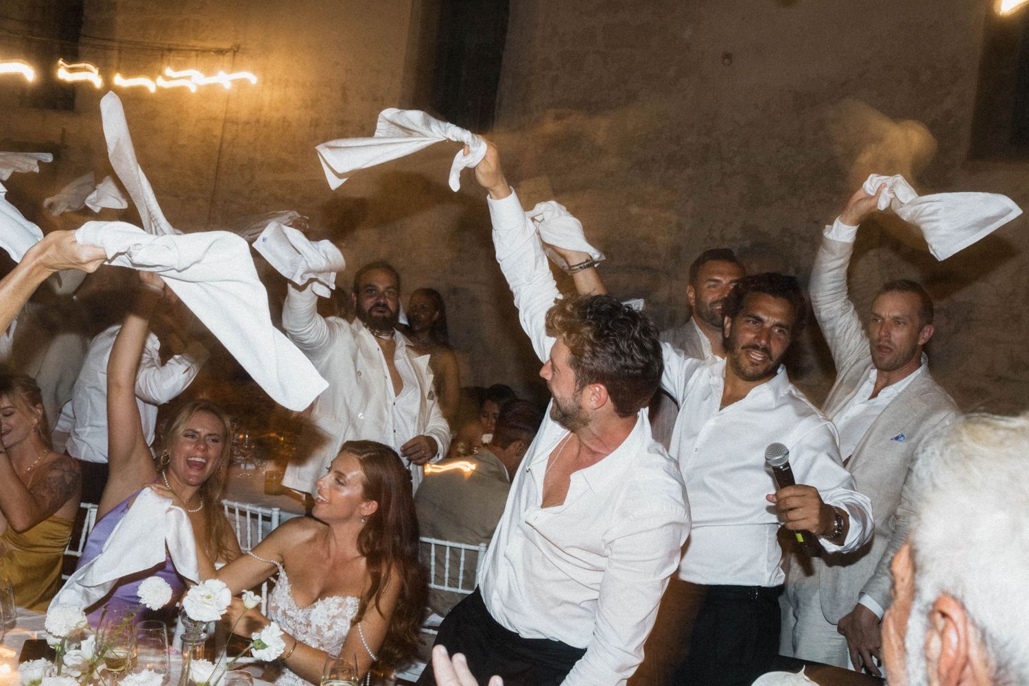 Wedding guests in white shirts wave napkins joyfully during lively indoor reception celebration.