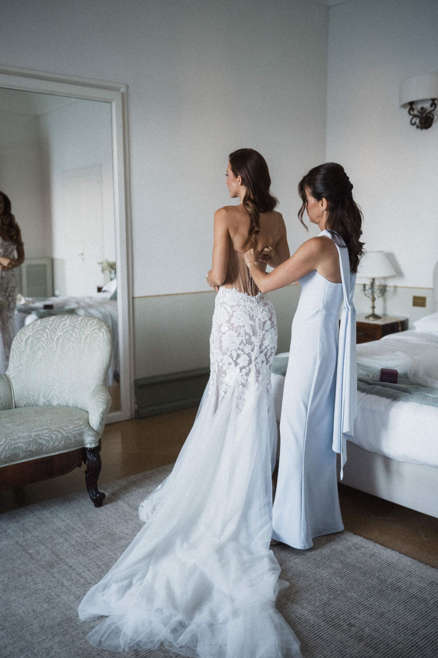 Bridesmaid in white dress fastening lace backless wedding gown for brunette bride reflected in elegant bedroom mirror.