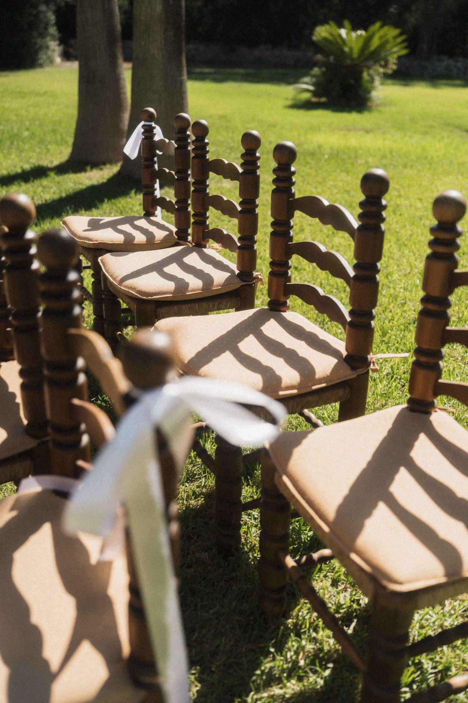 Elegant wooden ladder-back chairs with burlap cushions and white ribbon bows arranged on grass for a wedding.
