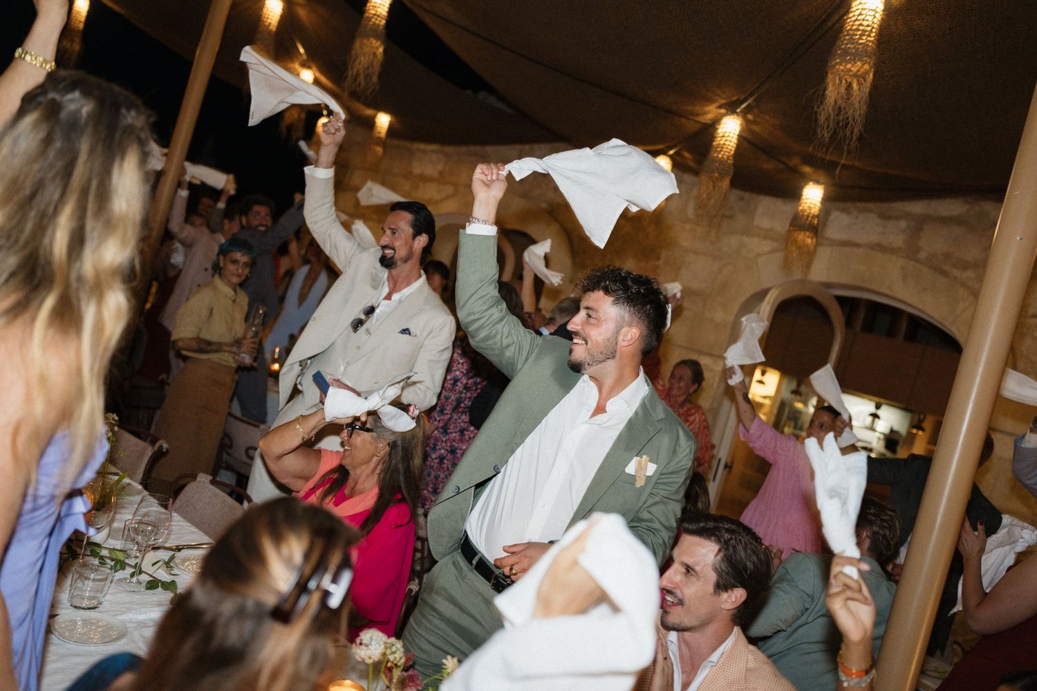Wedding guests wave white napkins and cheer during a lively evening reception under warm string lights.