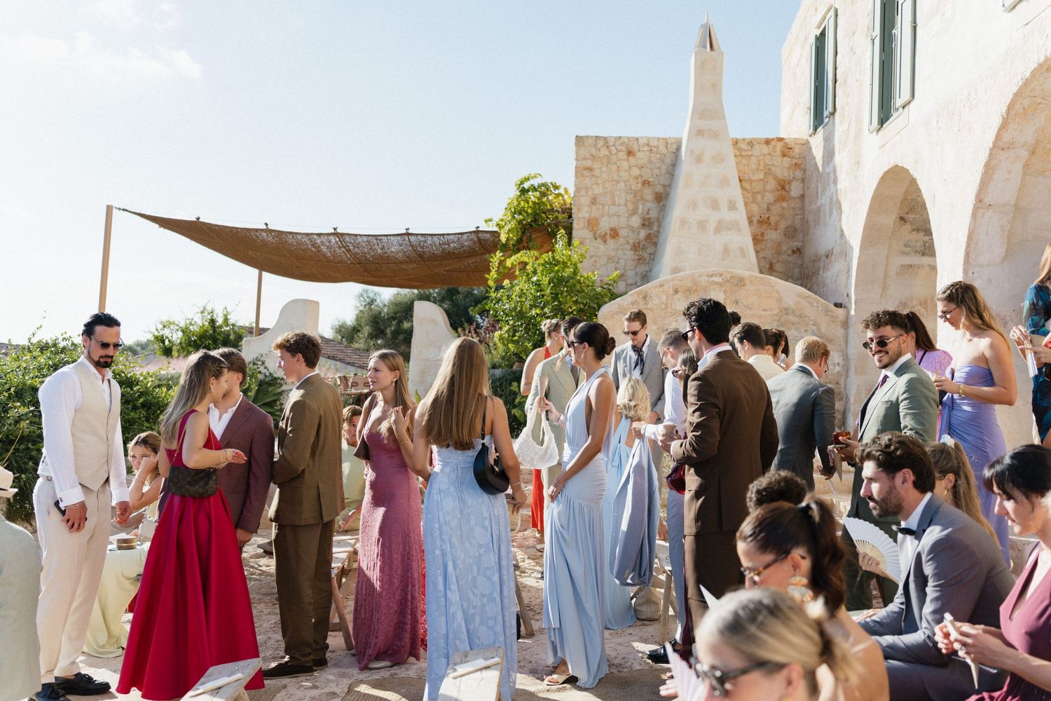 Wedding guests in colorful dresses and suits mingle on a sunny stone terrace beside a rustic Mediterranean chapel with a fabric canopy.
