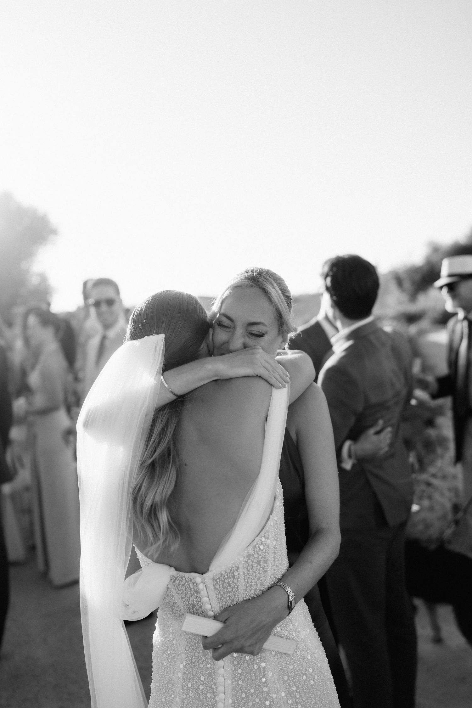 Black and white photo of the bride in a low-back beaded gown and veil sharing a joyful hug with a smiling guest.