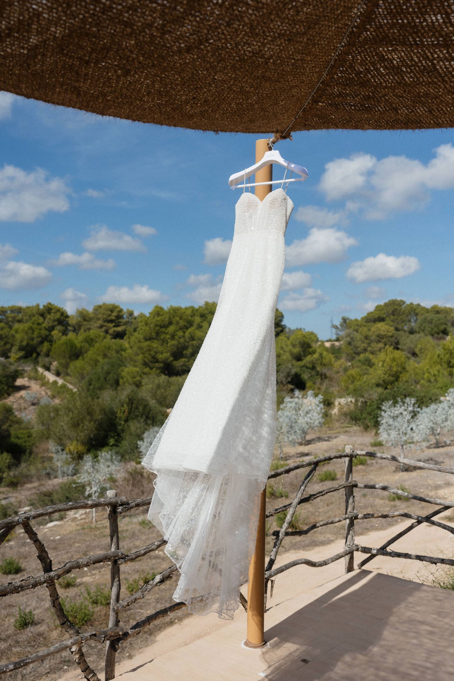 White lace wedding dress hanging on a white hanger from a wooden post under a thatched shade, with green hills and blue sky behind.