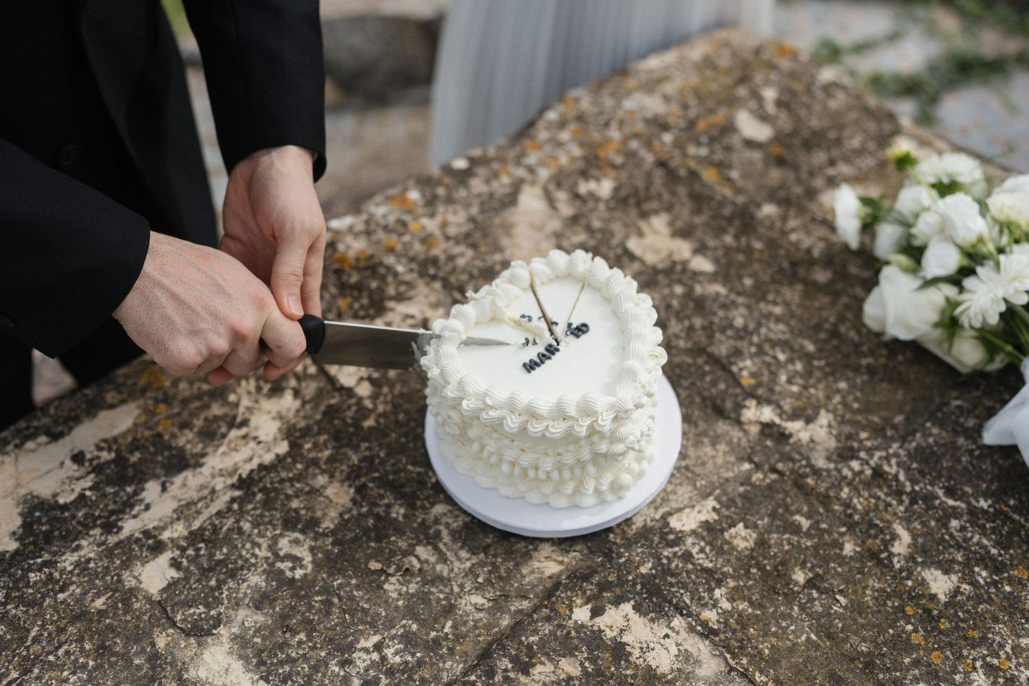 Person in black cutting a small white wedding cake with a knife on a rustic stone surface beside white flowers.