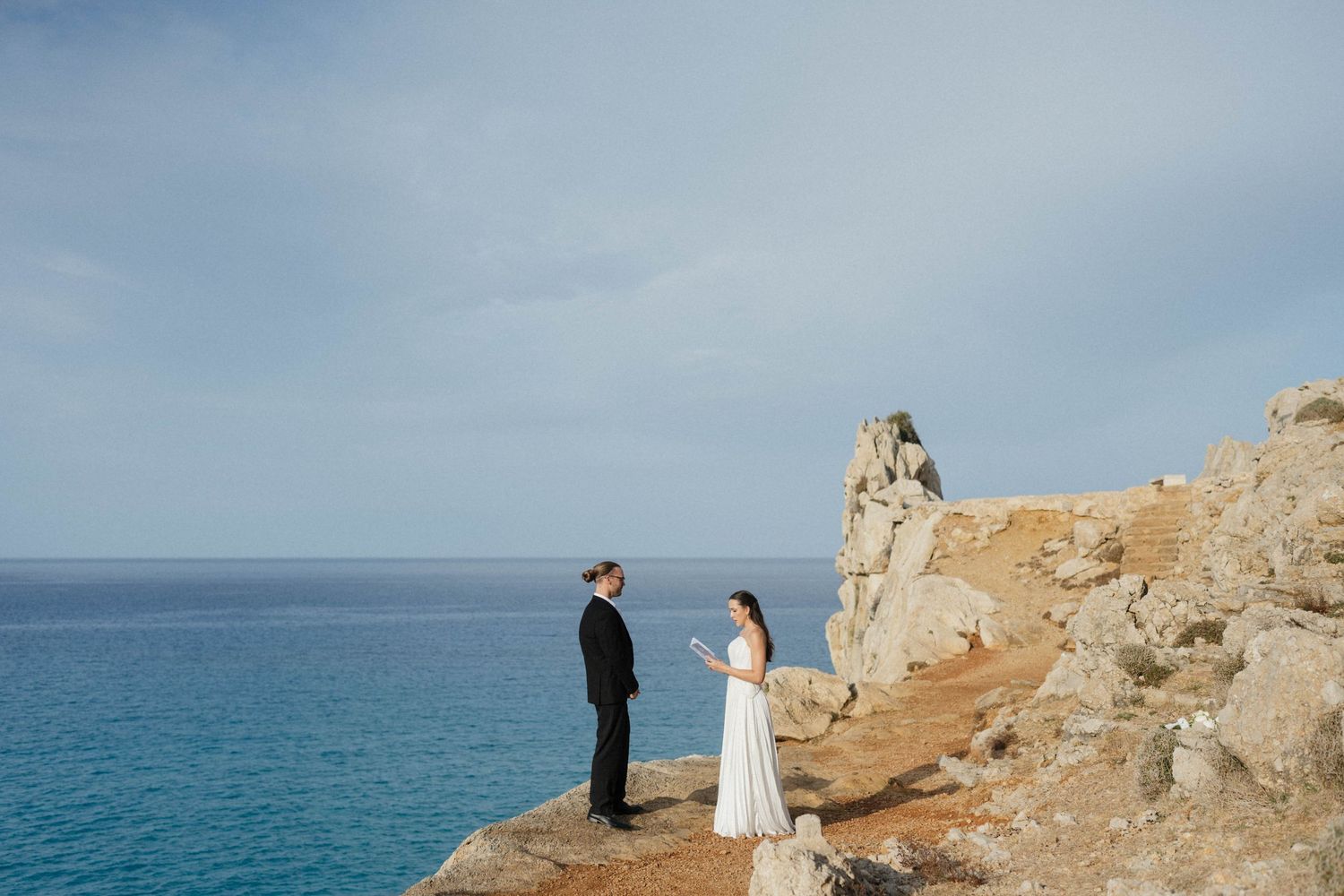 Bride in a white dress and groom in a dark suit stand facing each other on a rocky seaside cliff overlooking the blue ocean.