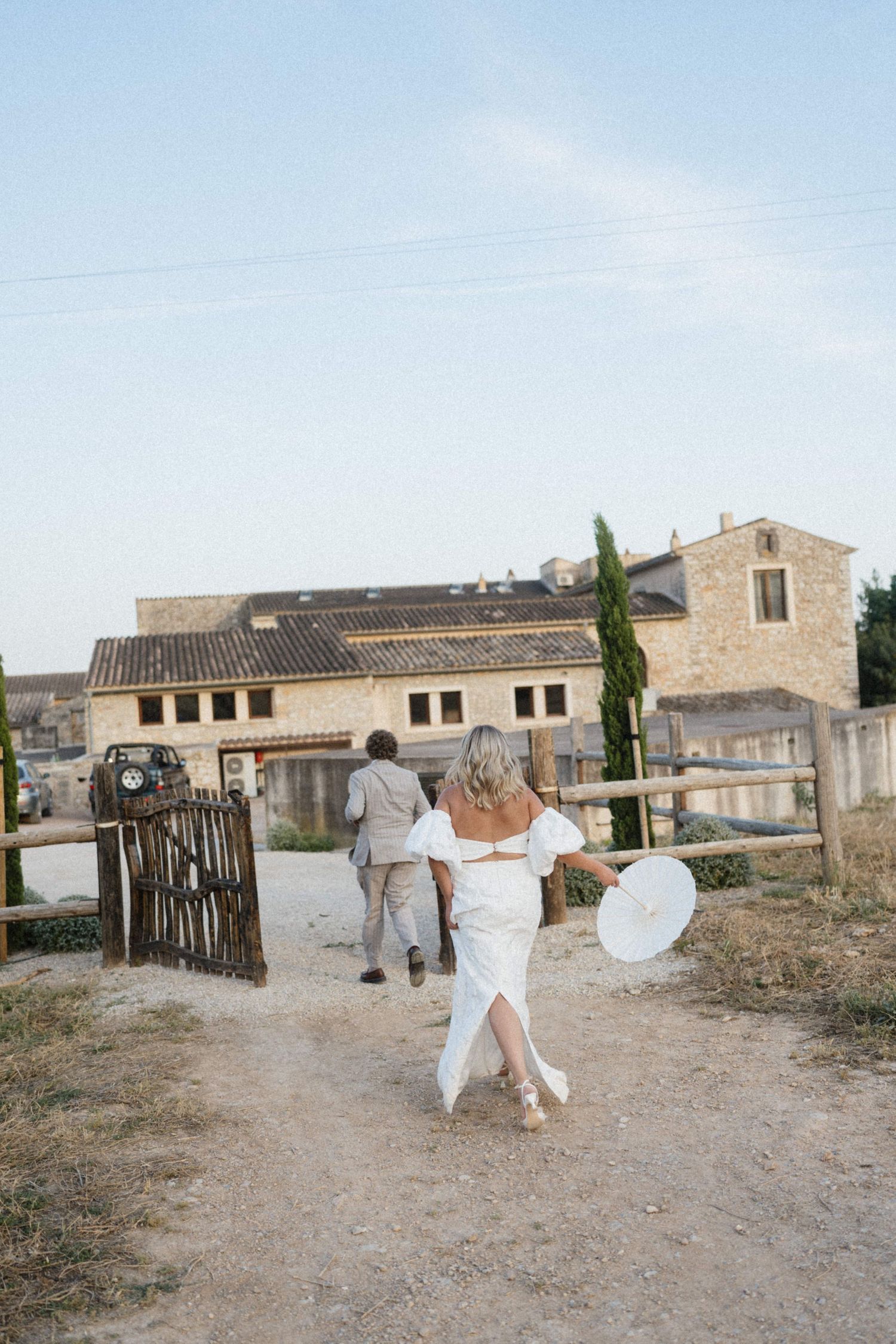 Bride in white off-shoulder gown walking toward rustic stone wedding venue.
