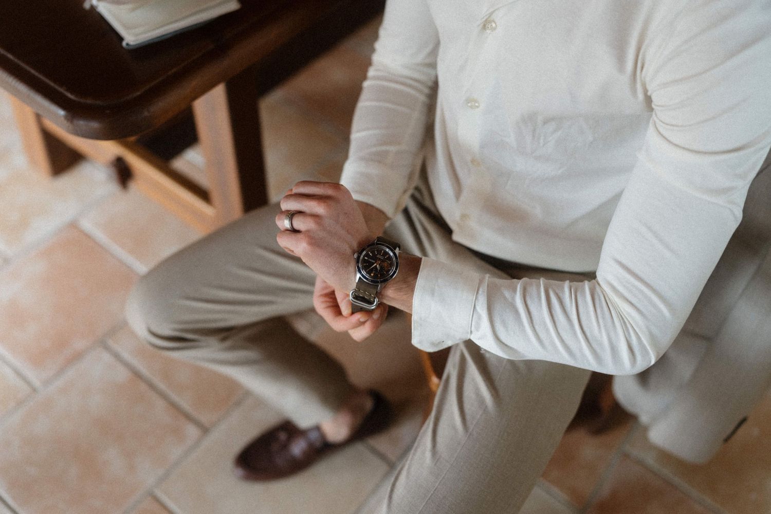 Man in white shirt and beige pants adjusting a dark wristwatch while seated, wearing brown leather loafers.