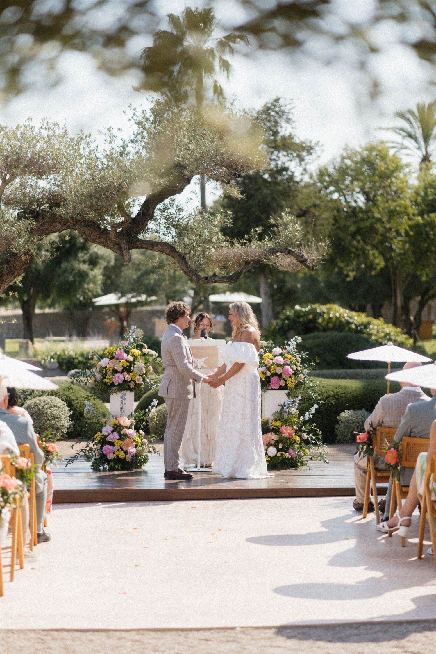 Bride and groom hold hands at outdoor altar surrounded by colorful floral arrangements and olive trees.
