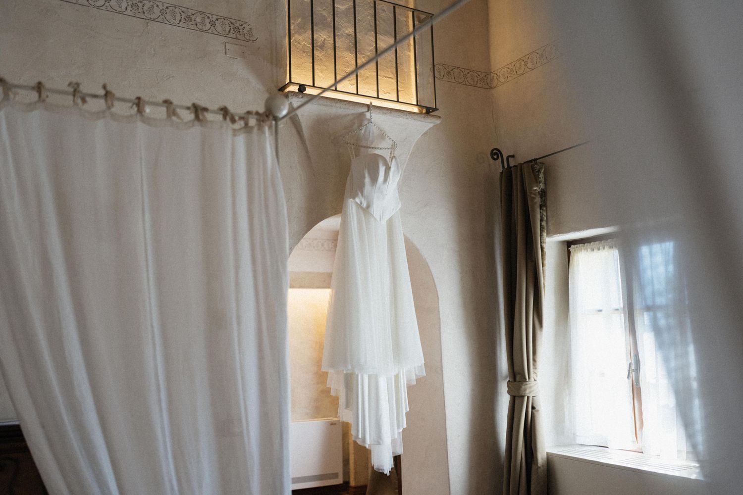 White wedding dress hanging from an ornate iron railing in an elegant bridal suite with curtains and natural light.