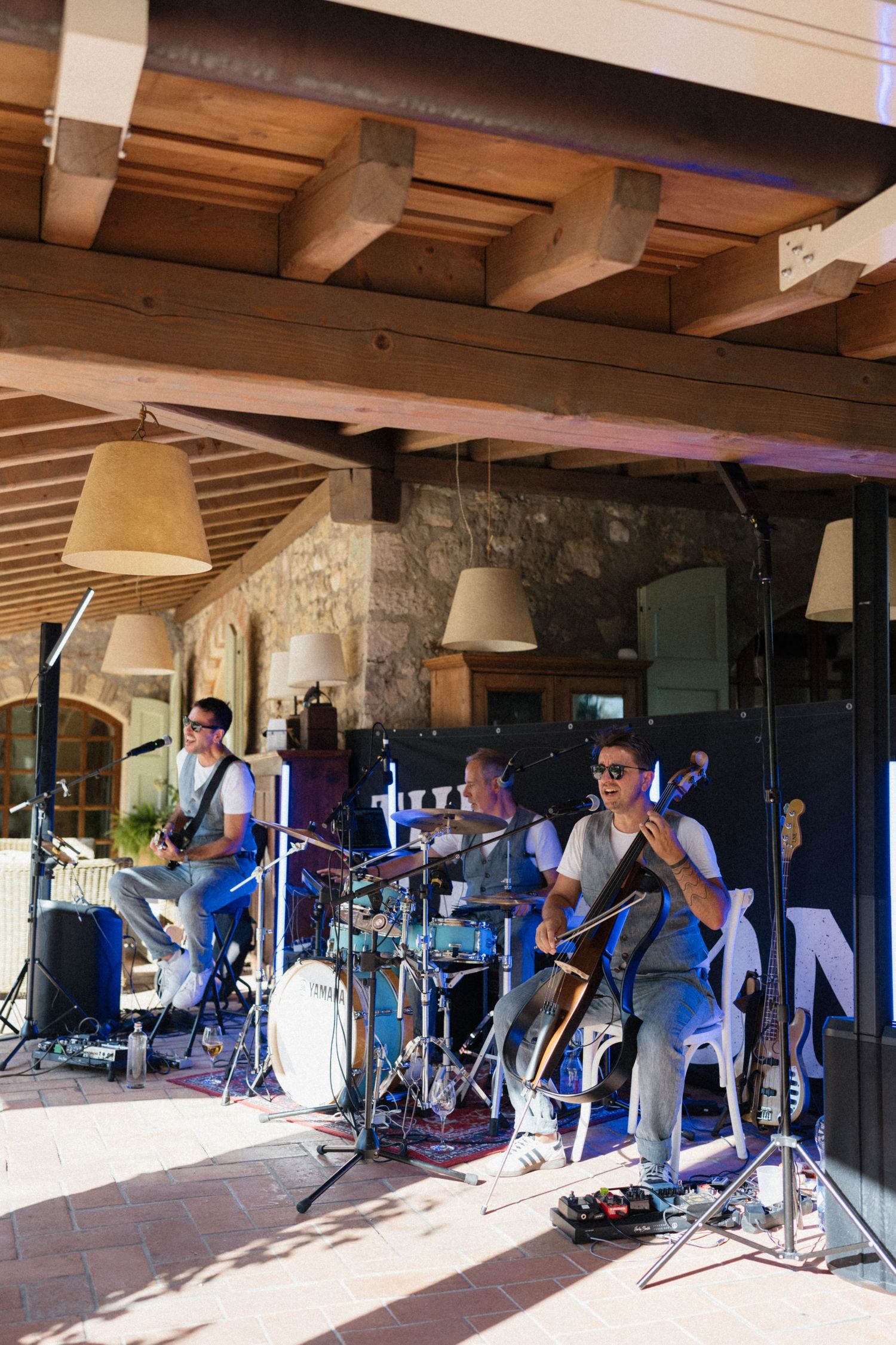 Live band performing outdoors under wooden beams, featuring guitar, bass, and drums at a rustic venue.