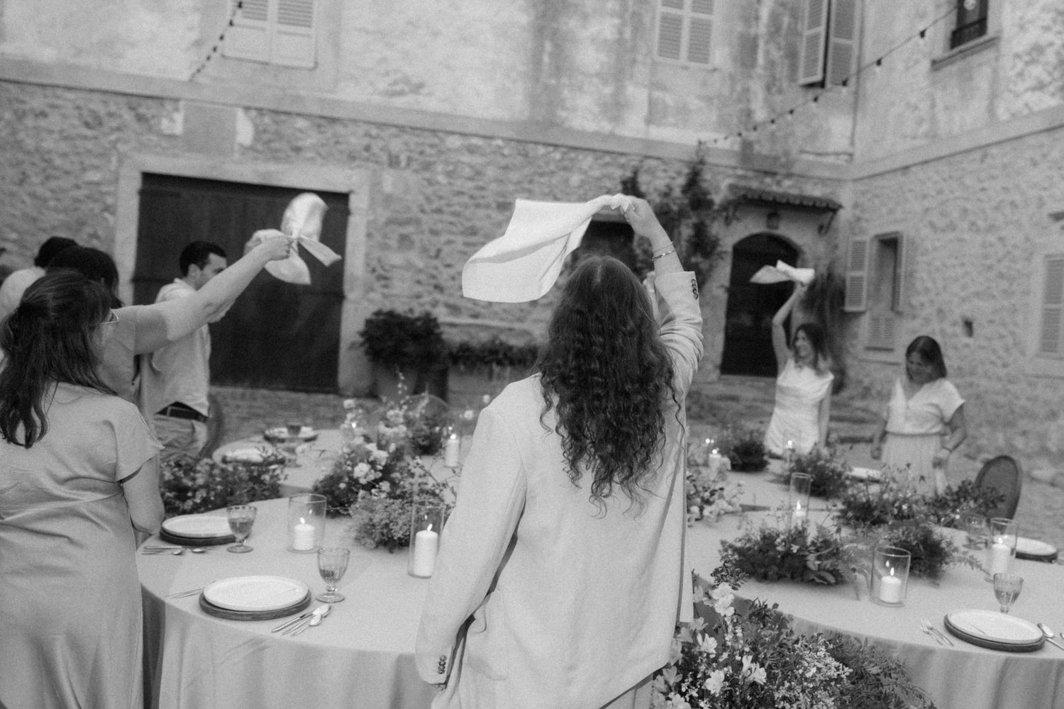 Black and white photo of wedding guests waving napkins over a floral decorated outdoor table.