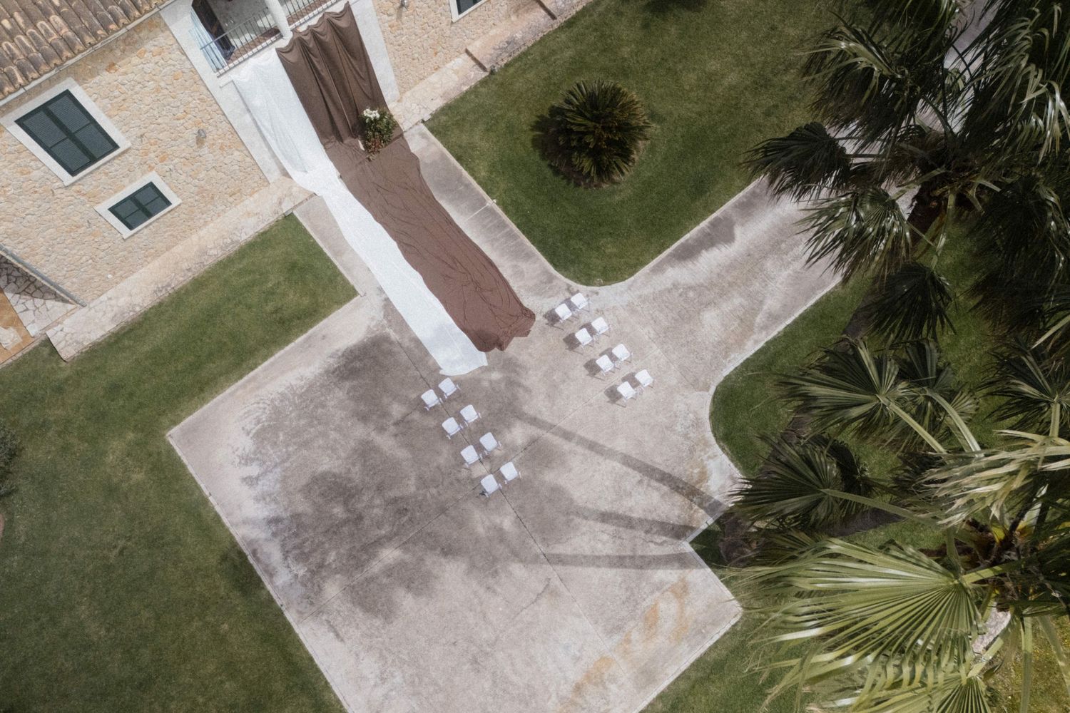 Aerial drone view of wedding venue with white chairs arranged outdoors among palm trees and green lawn.