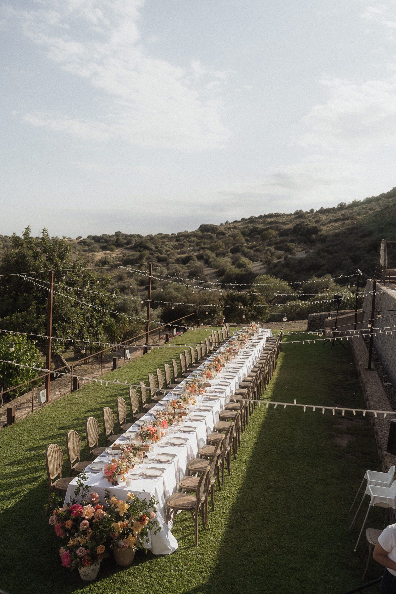 Elegant outdoor wedding reception with long banquet table, floral centerpieces, string lights, and lush green hills backdrop.