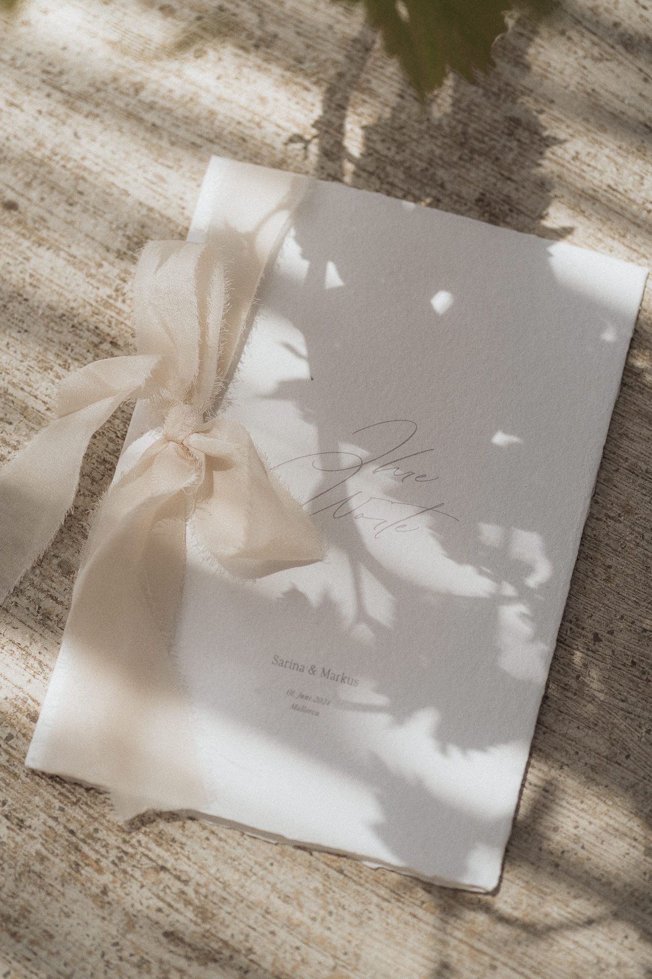 Elegant wedding program booklet with cream ribbon bow, featuring script typography, resting on textured surface with soft shadows.
