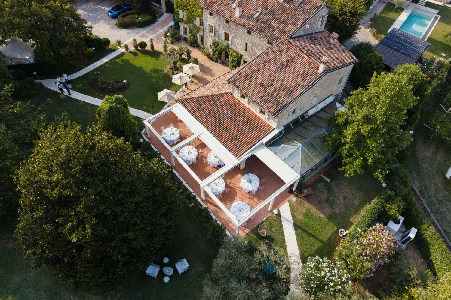 Aerial view of a luxury Italian villa with terracotta roof, rooftop terrace, swimming pool, and lush green gardens.