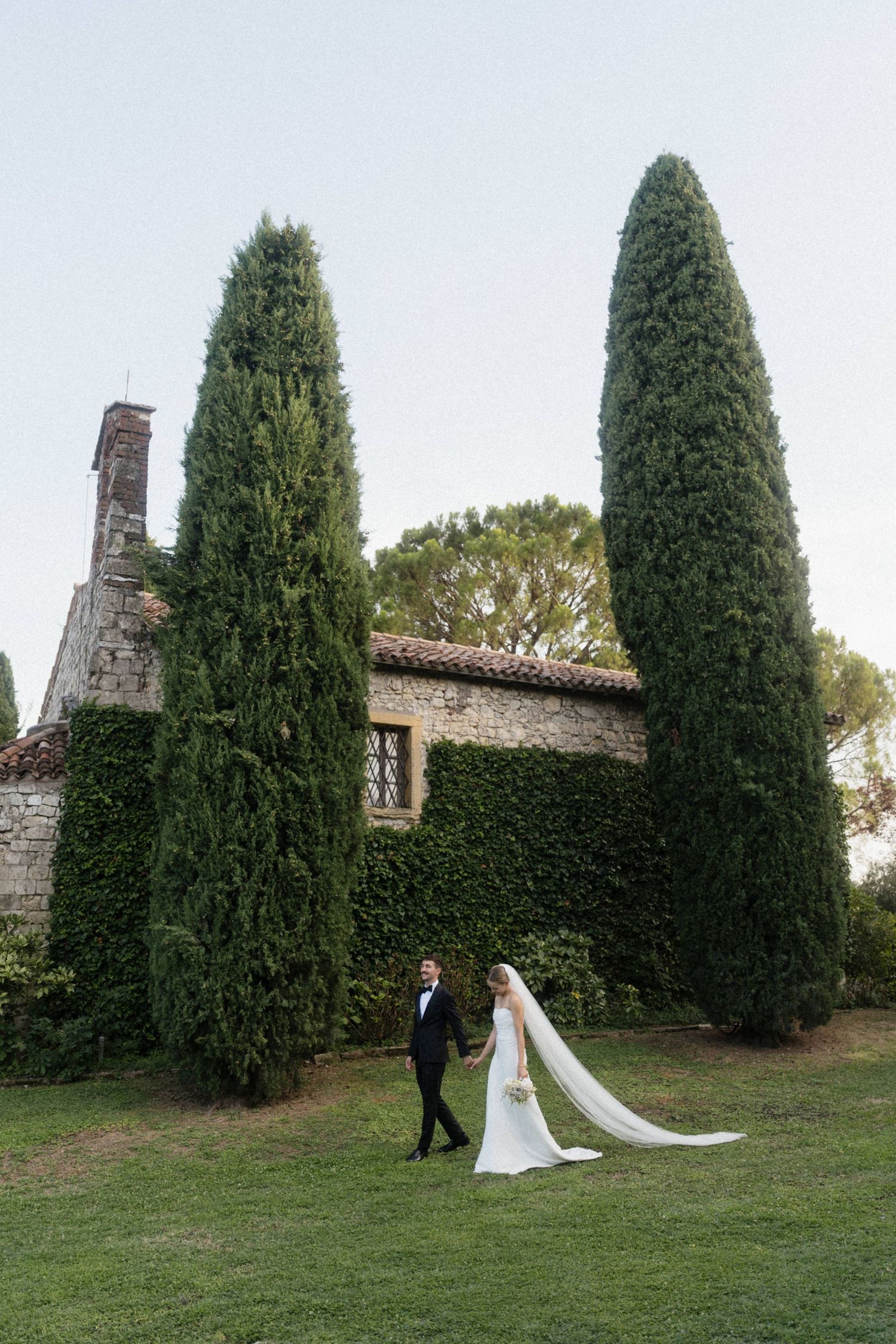 Bride and groom walk hand in hand on lush green lawn beside tall cypress trees and rustic stone villa in Tuscany.