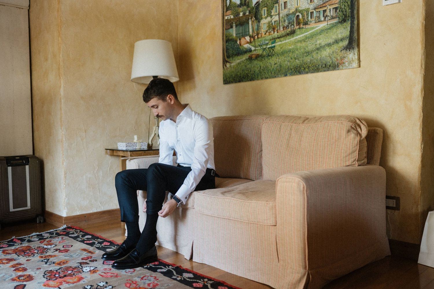 Groom in white shirt and black pants sitting on beige sofa adjusting shoes in elegant room.