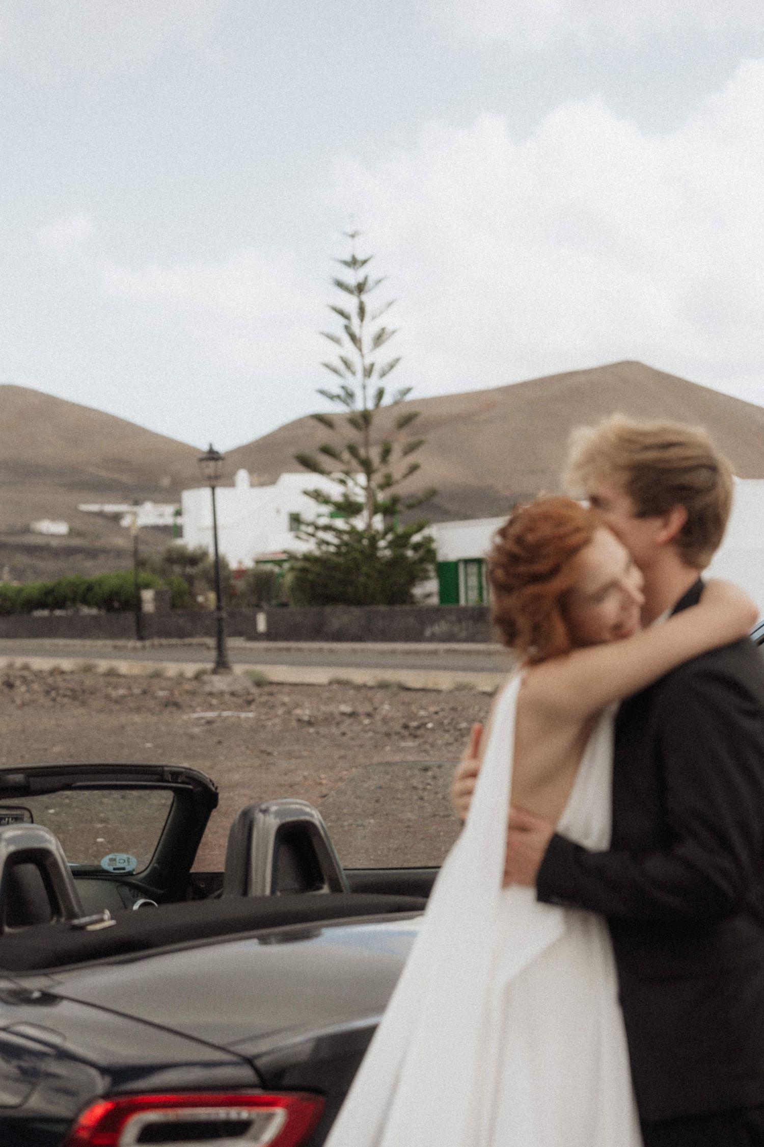 Bride and groom embracing near convertible car with volcanic mountain landscape behind them.