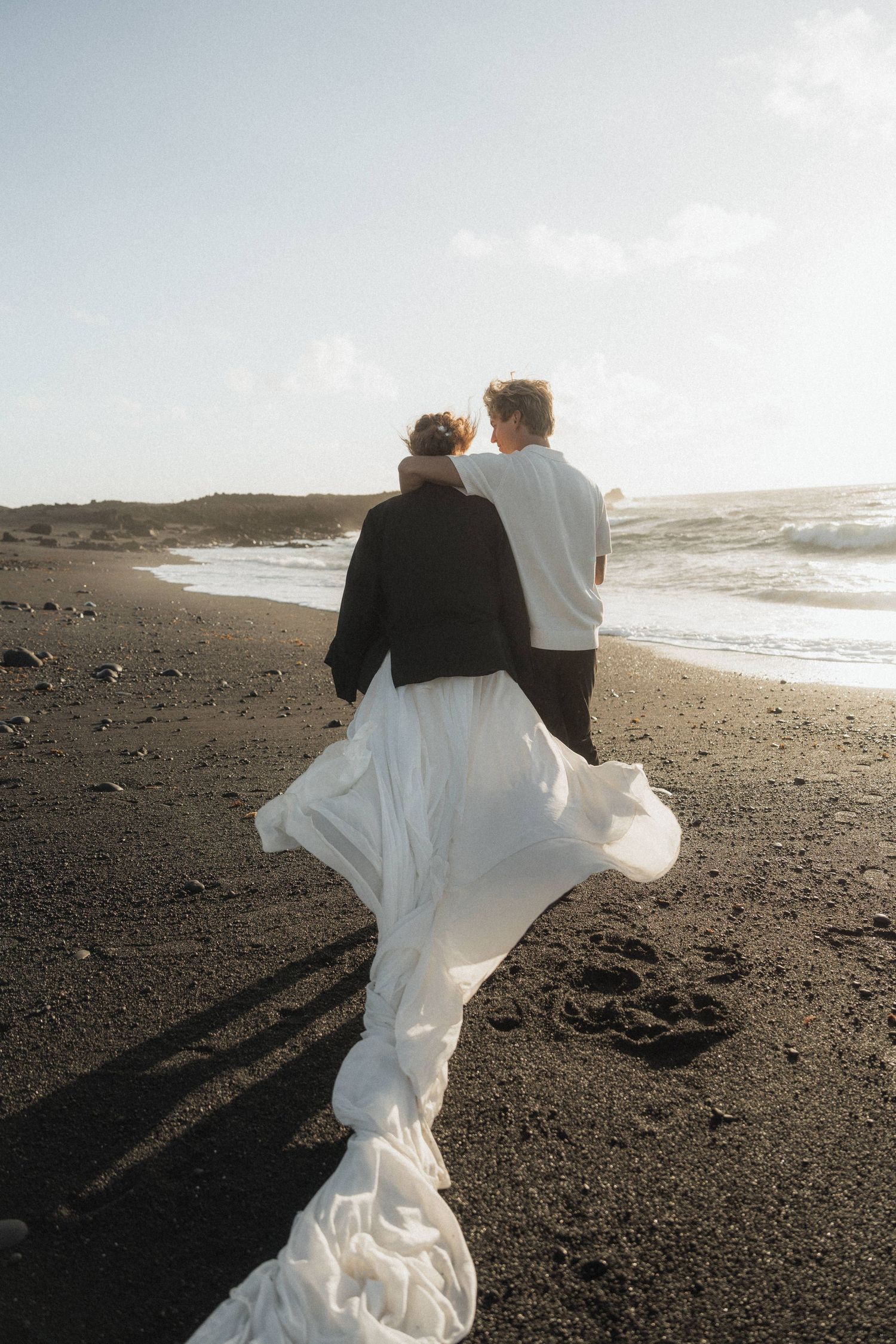 Couple in wedding attire walking on black sand beach, bride's white dress flowing in wind.