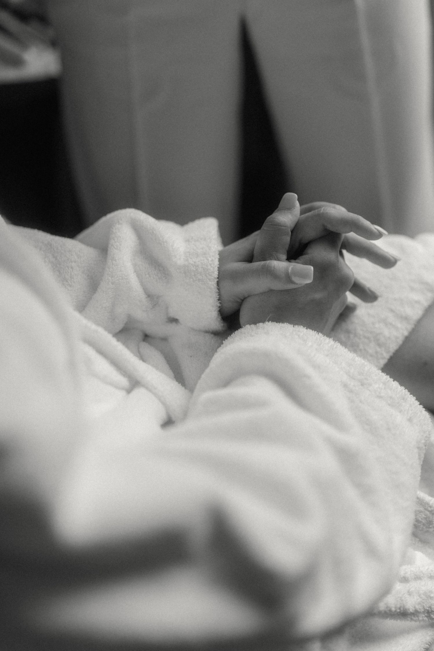 Black and white close-up of bride's hands clasped in white fluffy robe.