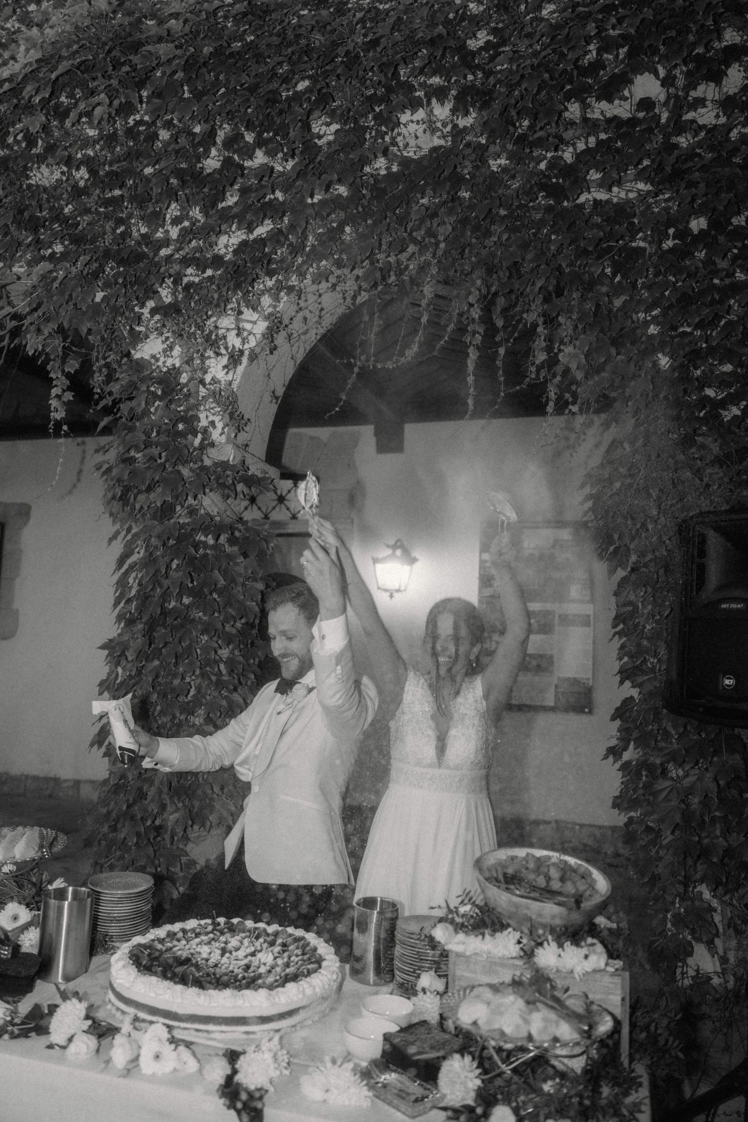Black and white photo of joyful bride and groom celebrating at their wedding cake table outdoors.