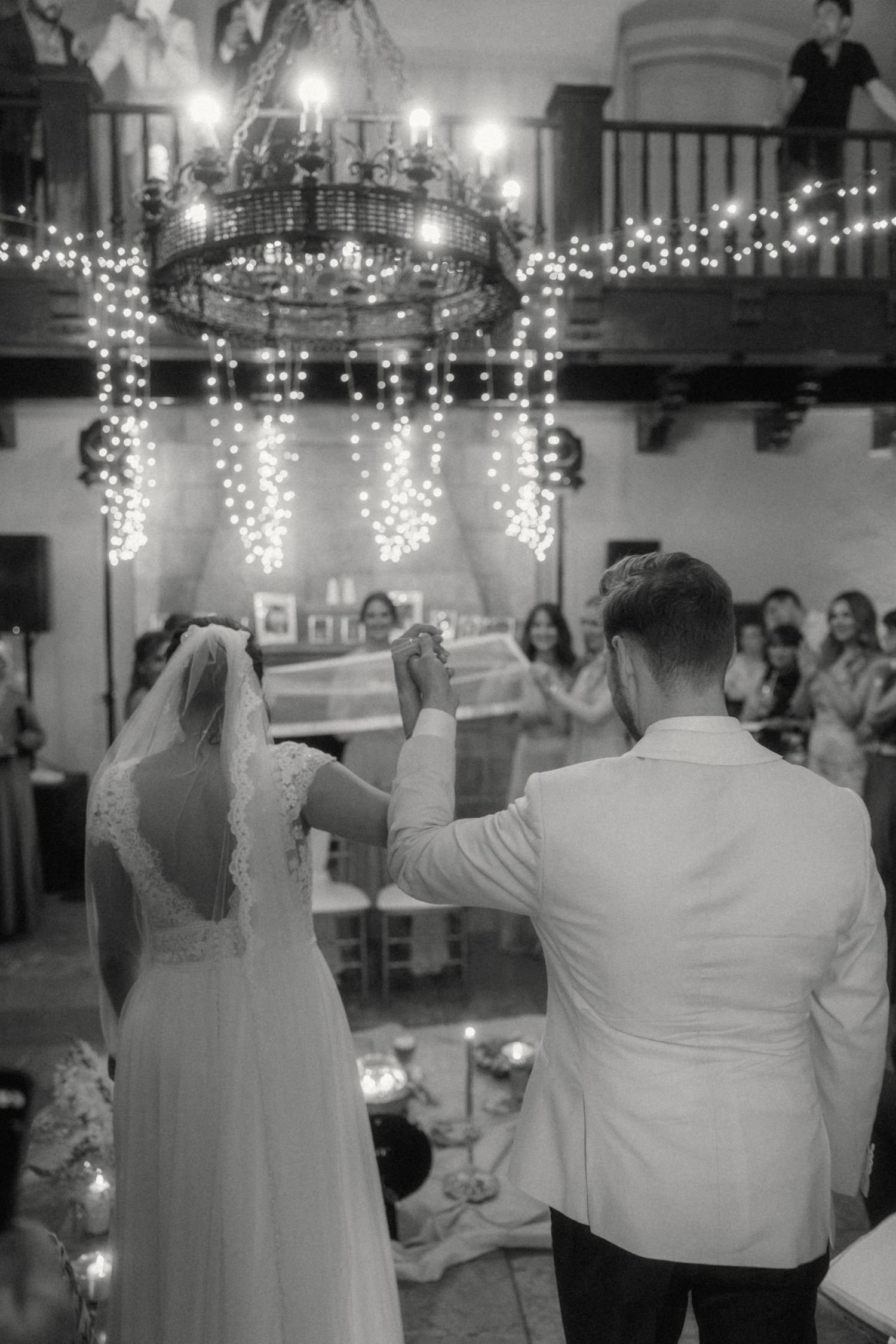 Black and white photo of bride and groom entering wedding reception under chandelier with fairy lights.