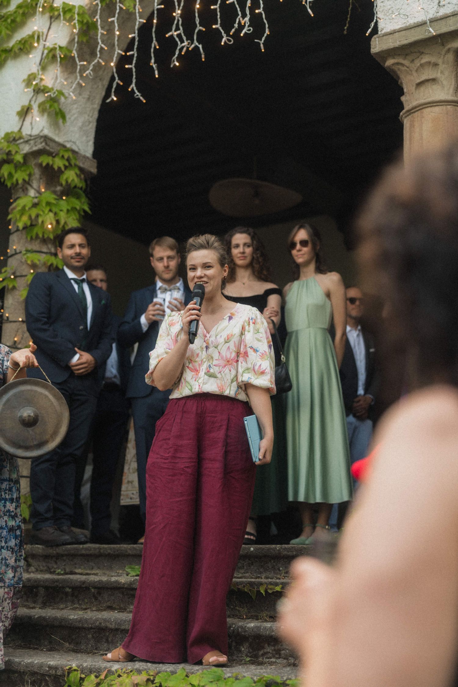 Woman in floral top and burgundy pants speaks into microphone on stone steps at outdoor wedding ceremony.