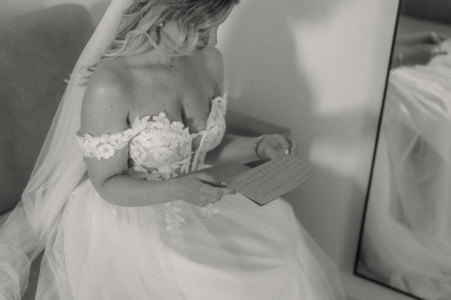 Bride in off-shoulder lace wedding gown reading a letter while seated near a mirror, black and white photo.