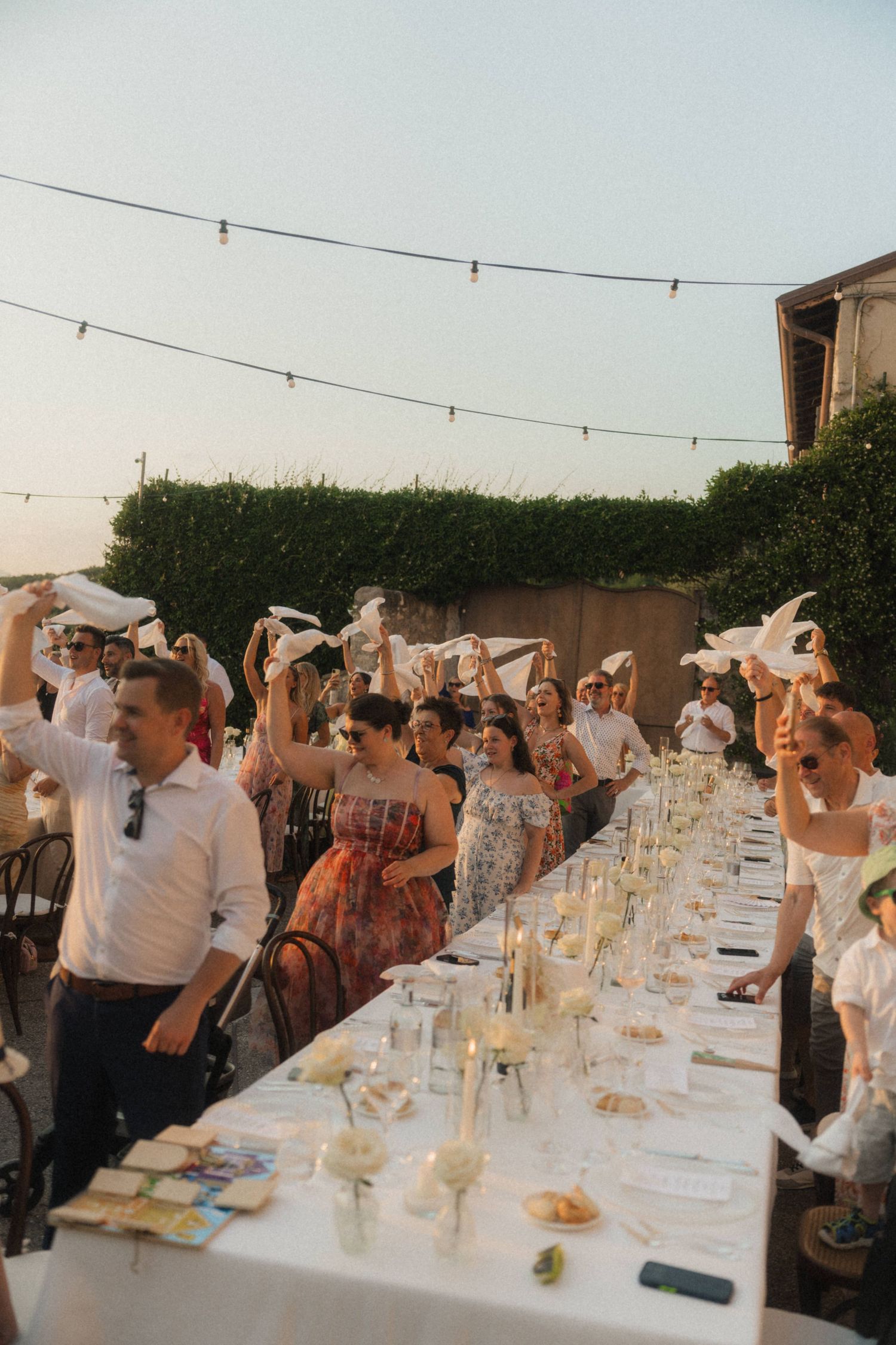 Wedding guests waving white napkins in celebration at outdoor candlelit reception dinner.
