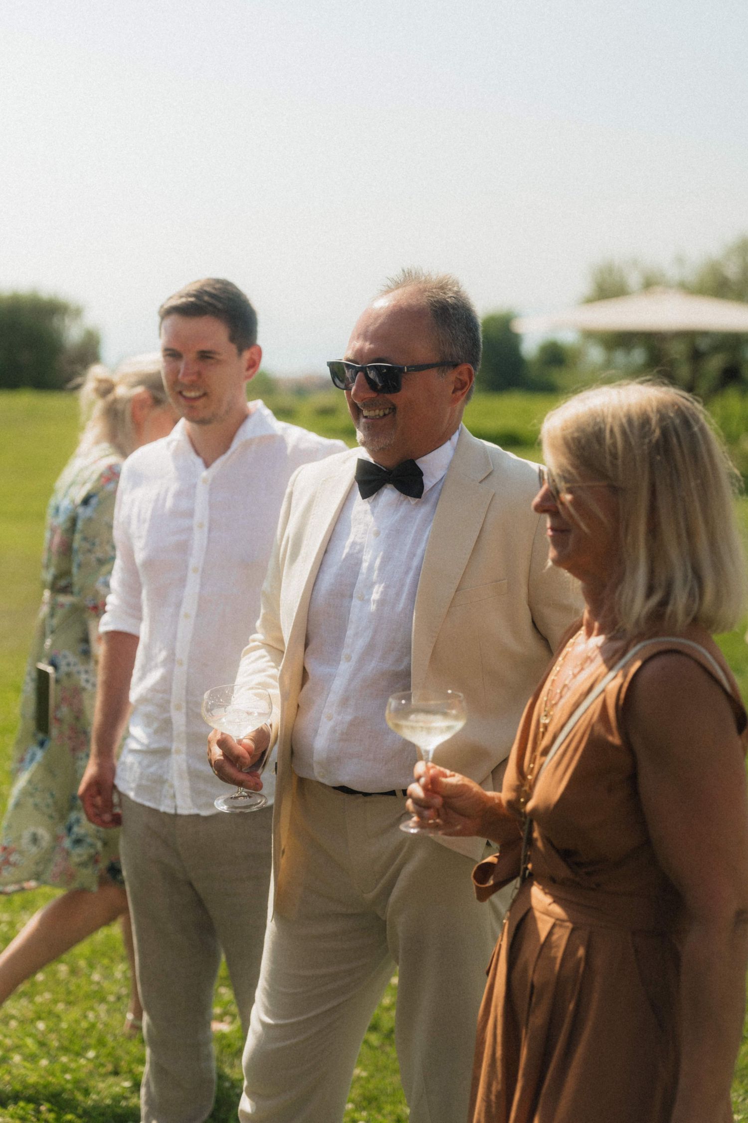 Elegant wedding guests in summer attire socialize outdoors holding wine glasses on sunny day.