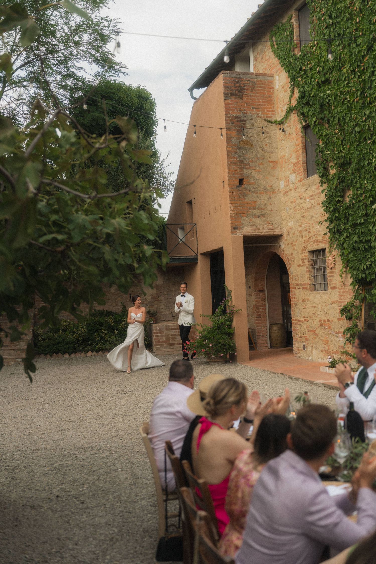 Bride and groom stand before a rustic Tuscan villa while wedding guests applaud at an outdoor reception dinner.